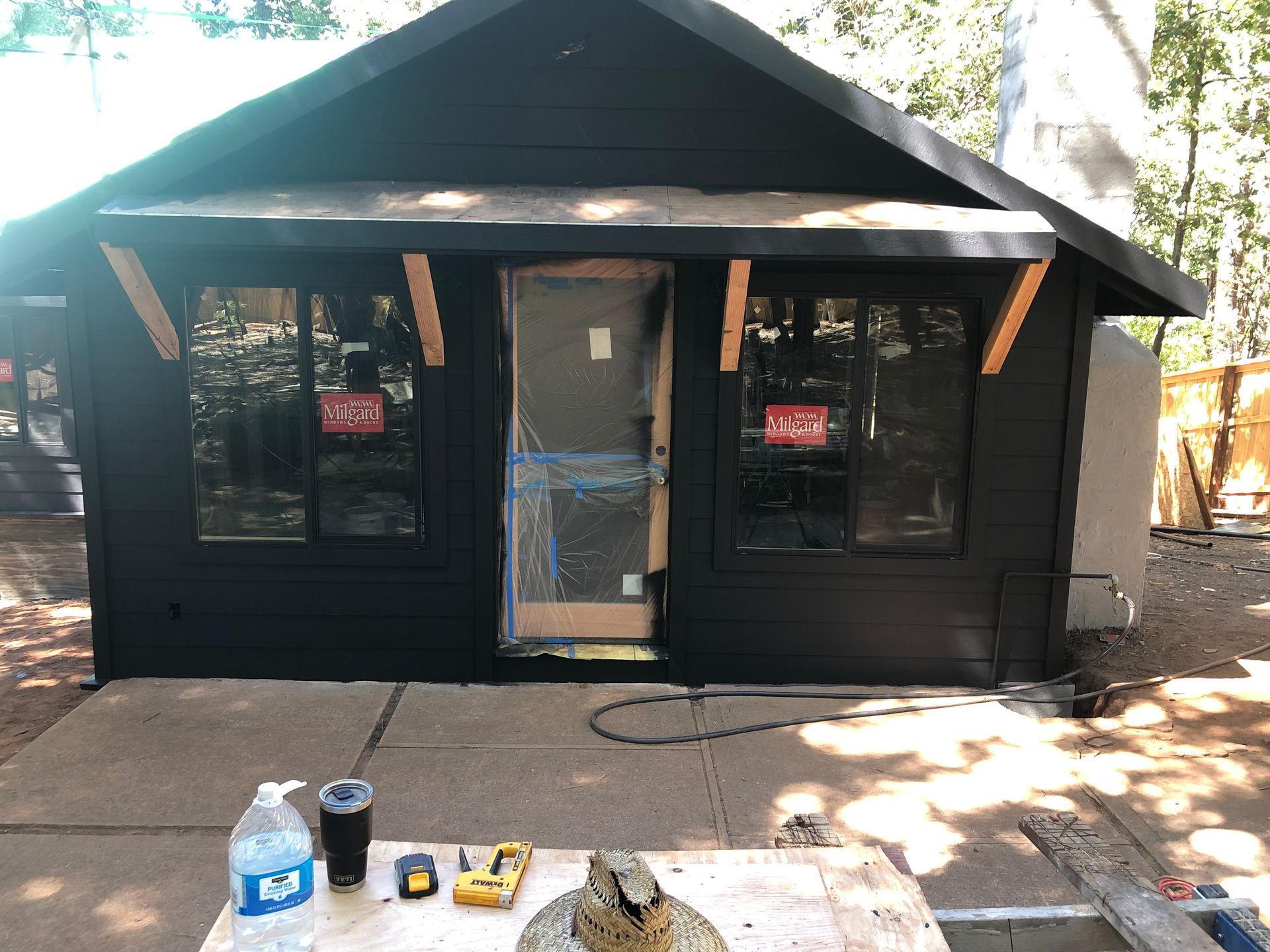 A black cabin exterior with a centered door and two windows under a small roof overhang, viewed from a patio.