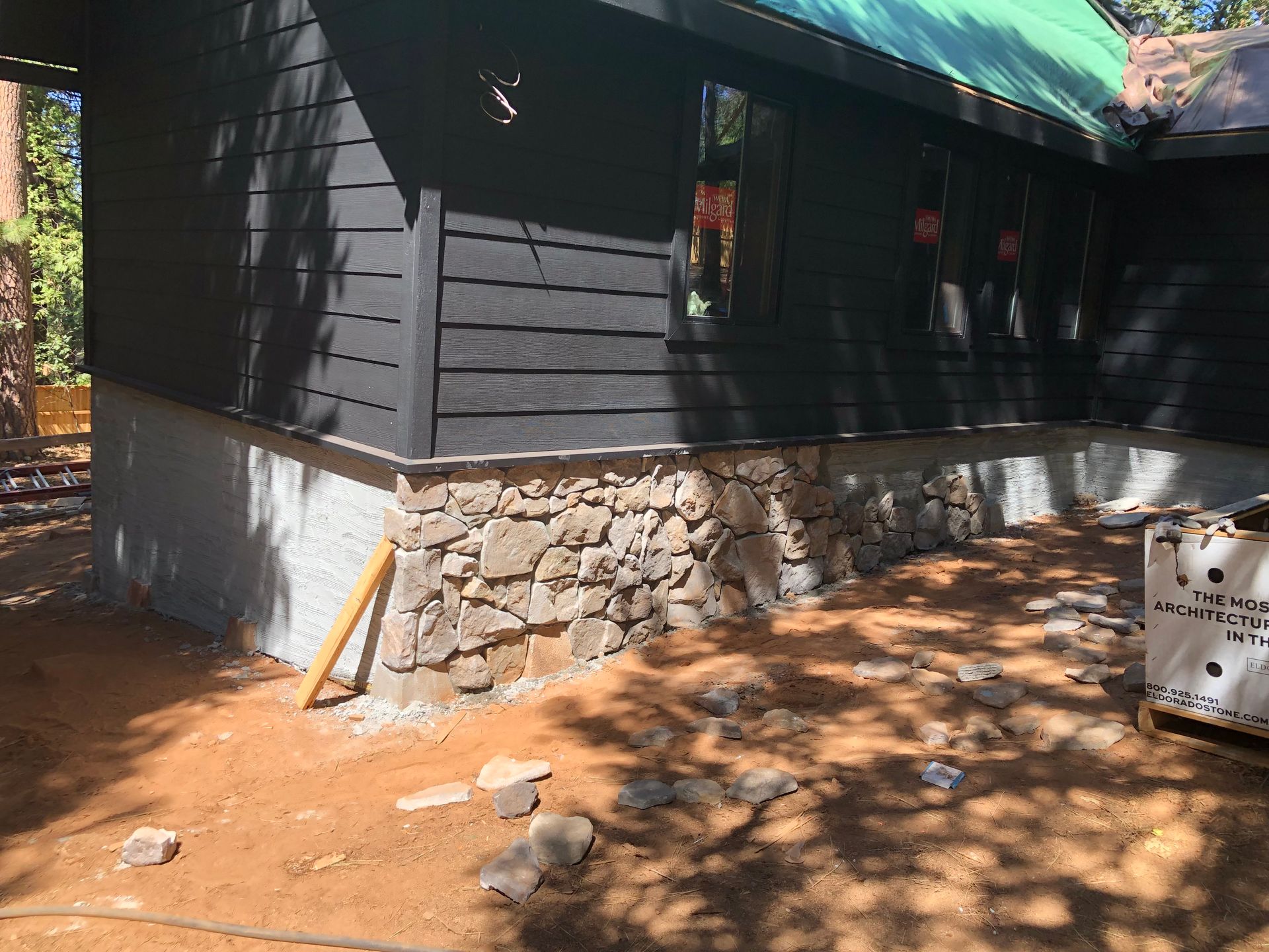 Exterior view of a home with black wood siding and a base of decorative stone on a dirt lot with a green roof.