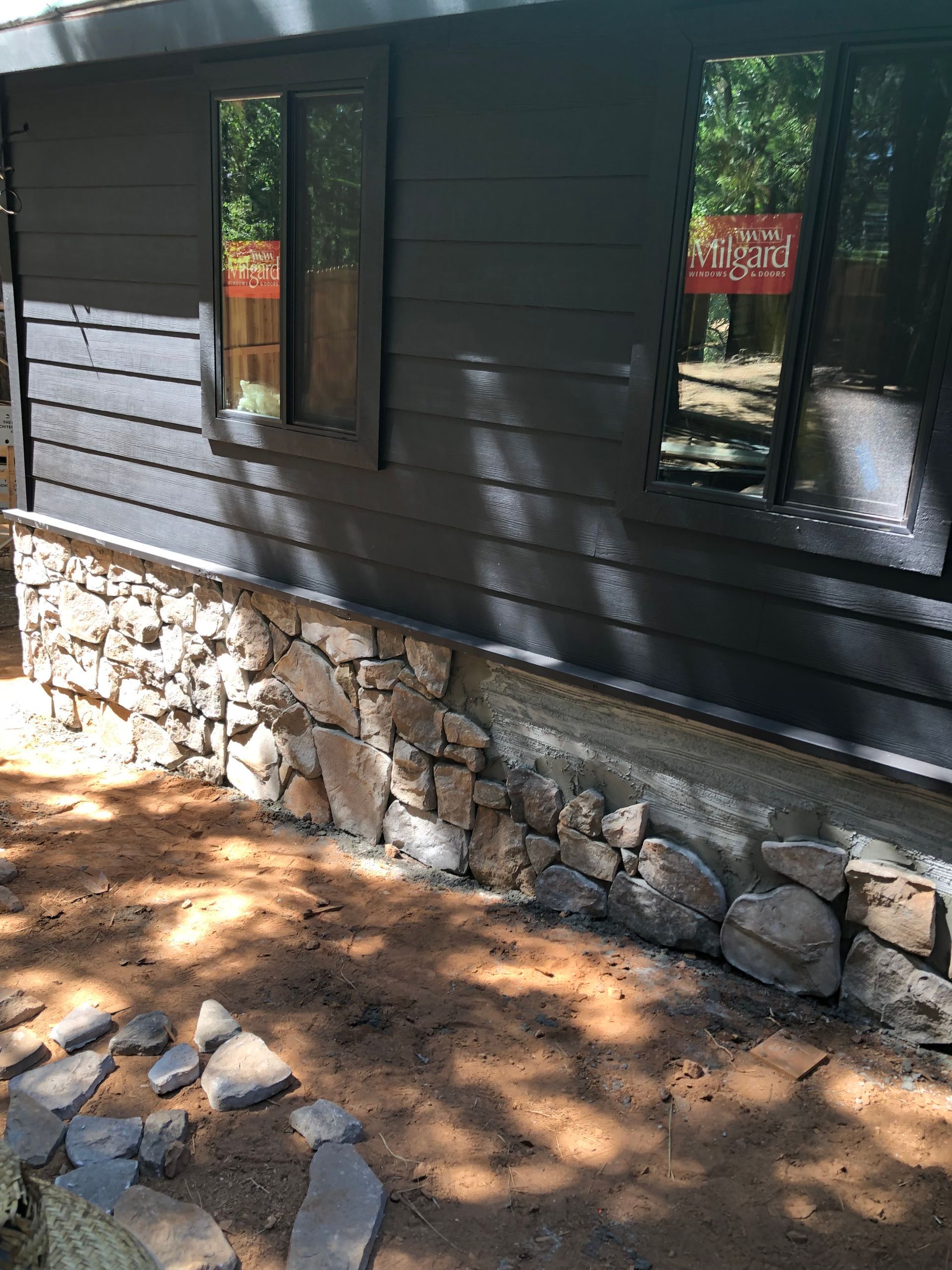 Exterior wall of a dark-painted building with horizontal siding above a stone foundation base, set in a dirt yard.