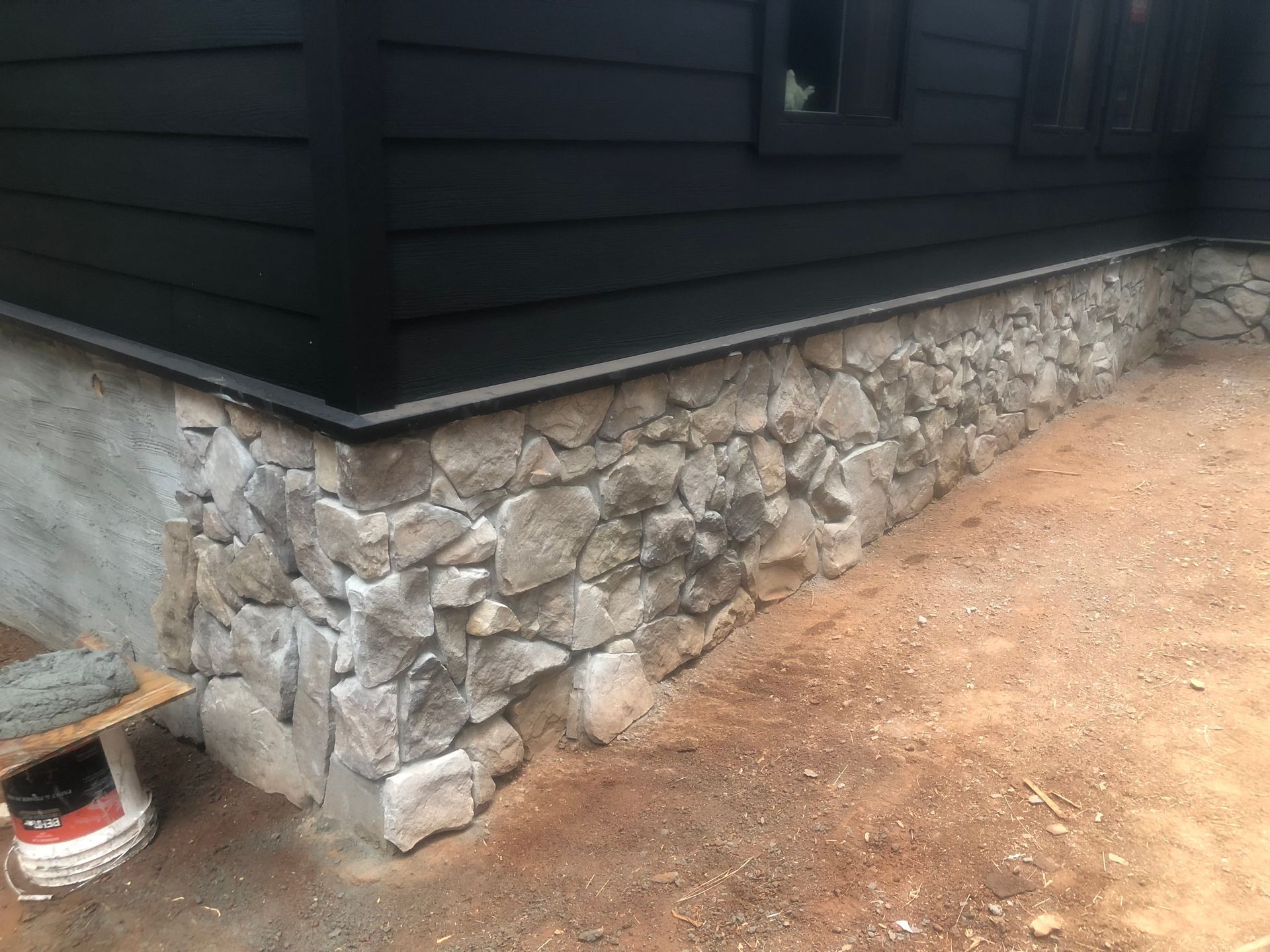 A house exterior featuring a foundation wall of light-colored, irregular stone veneer below black horizontal siding.
