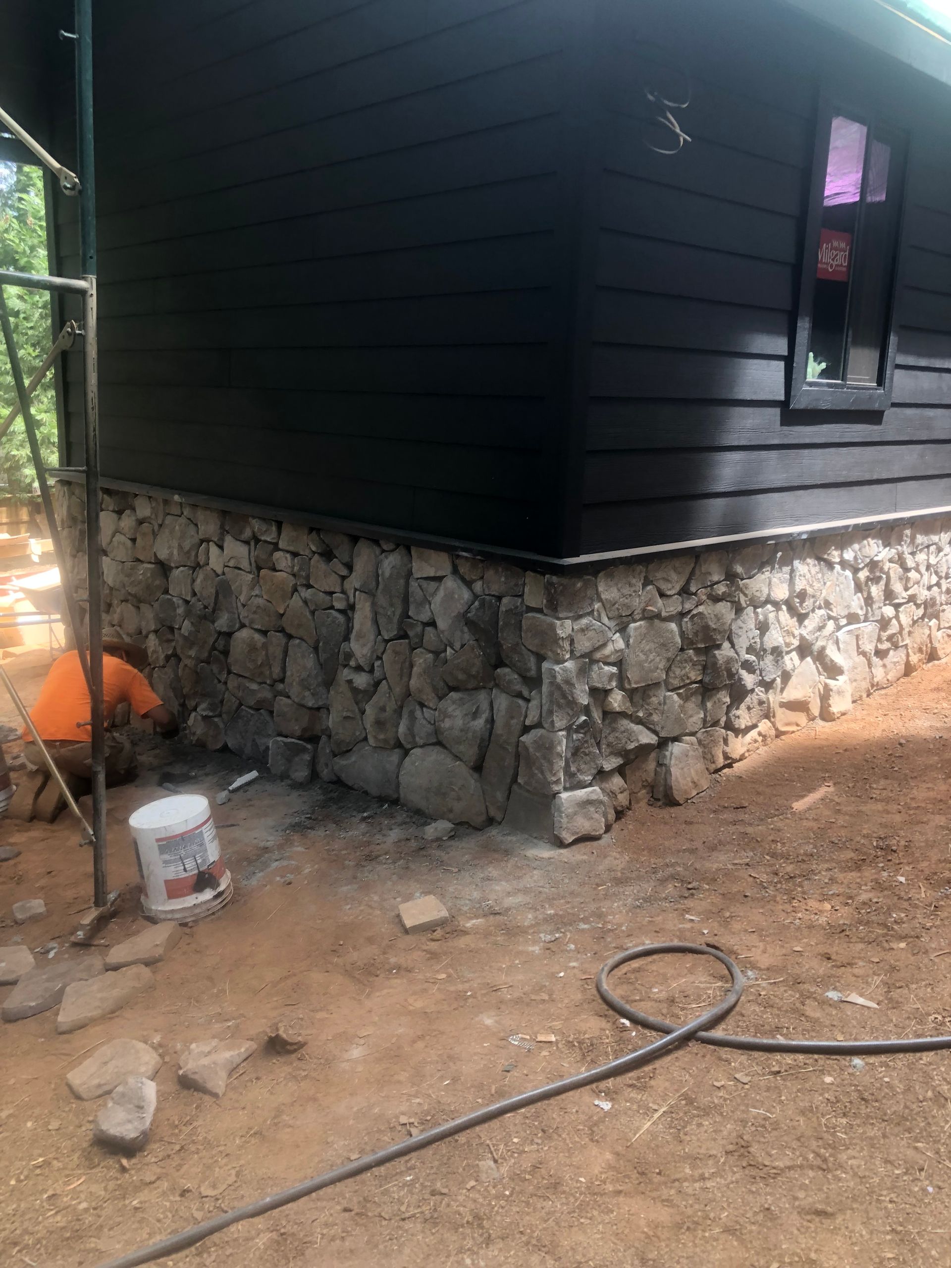 A worker installing a natural stone foundation on the exterior of a black-sided building.