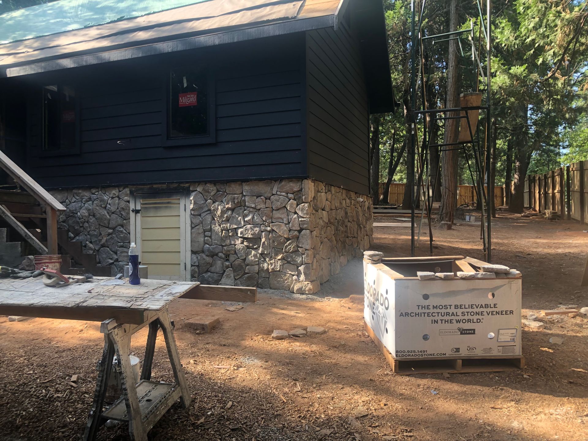 A dark-sided cabin with a stone foundation under construction, featuring a wooden table and supply crate in a yard.