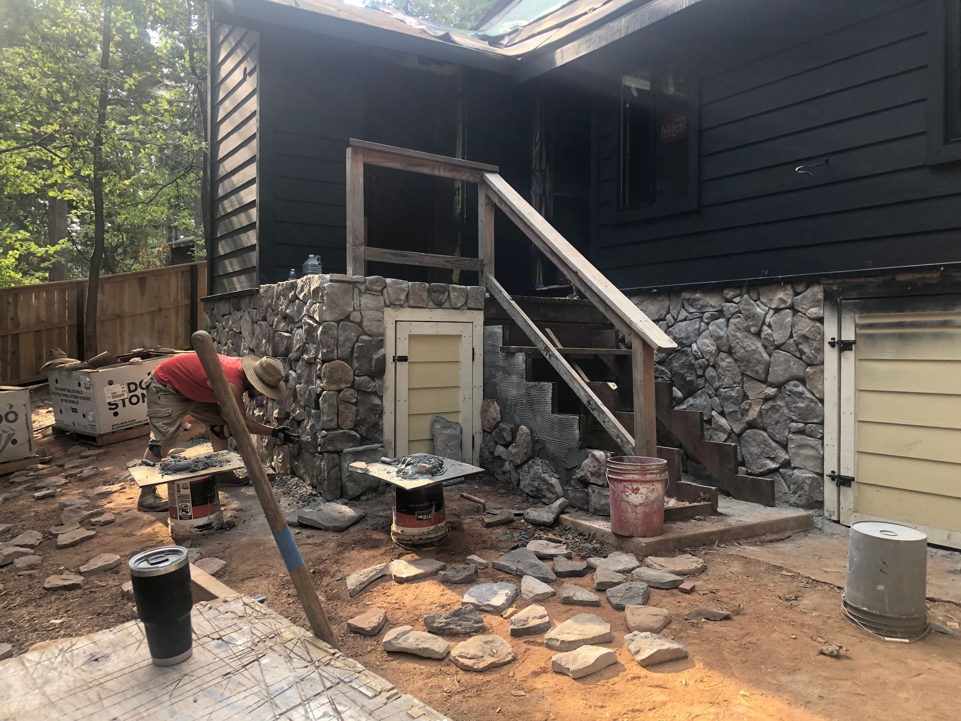 A person in a red shirt works on a stone wall exterior project beneath a dark-sided house with wooden steps.