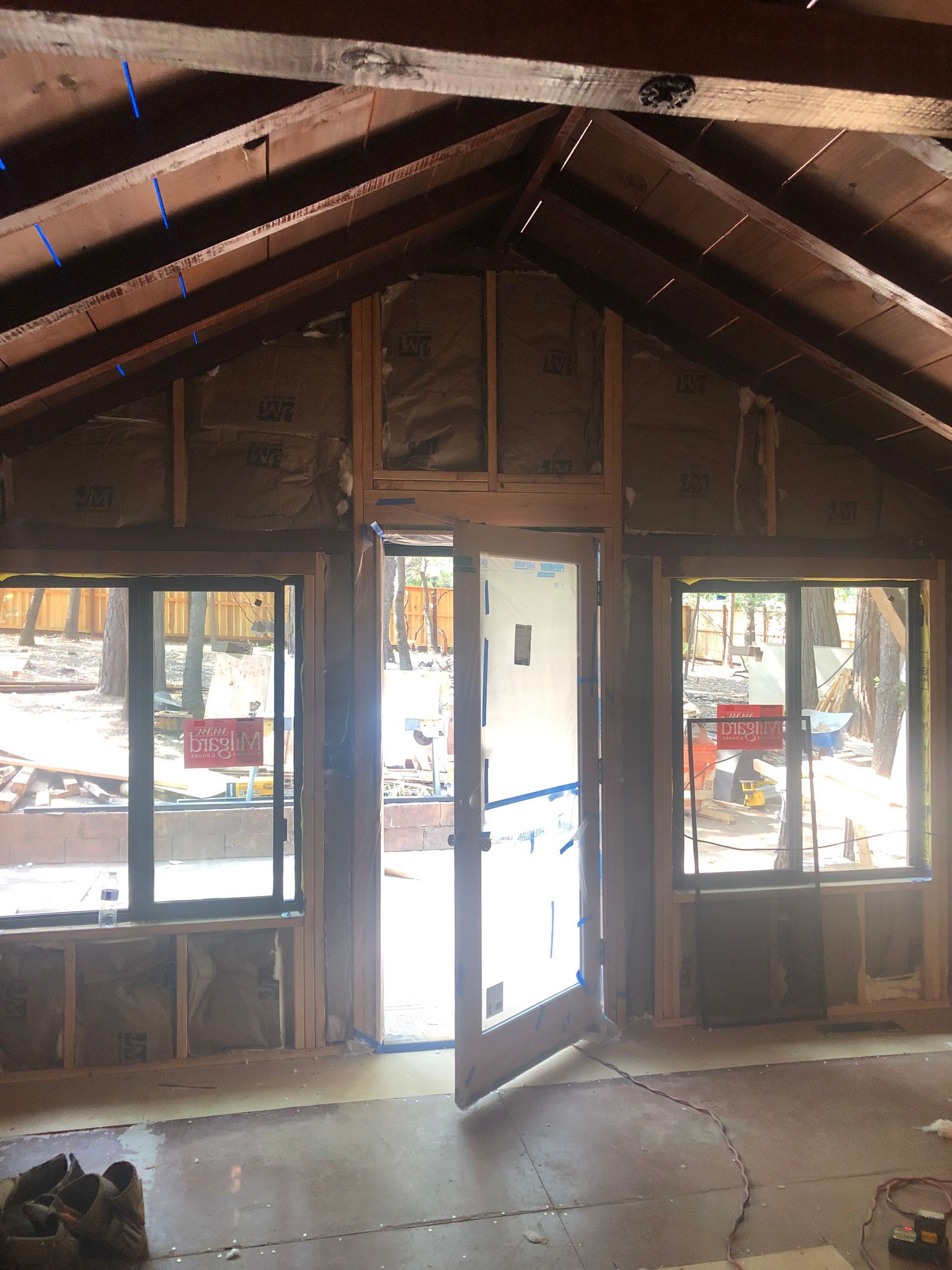 Unfinished interior room with exposed wooden beams, insulation between wall studs, two windows, and an open glass door.