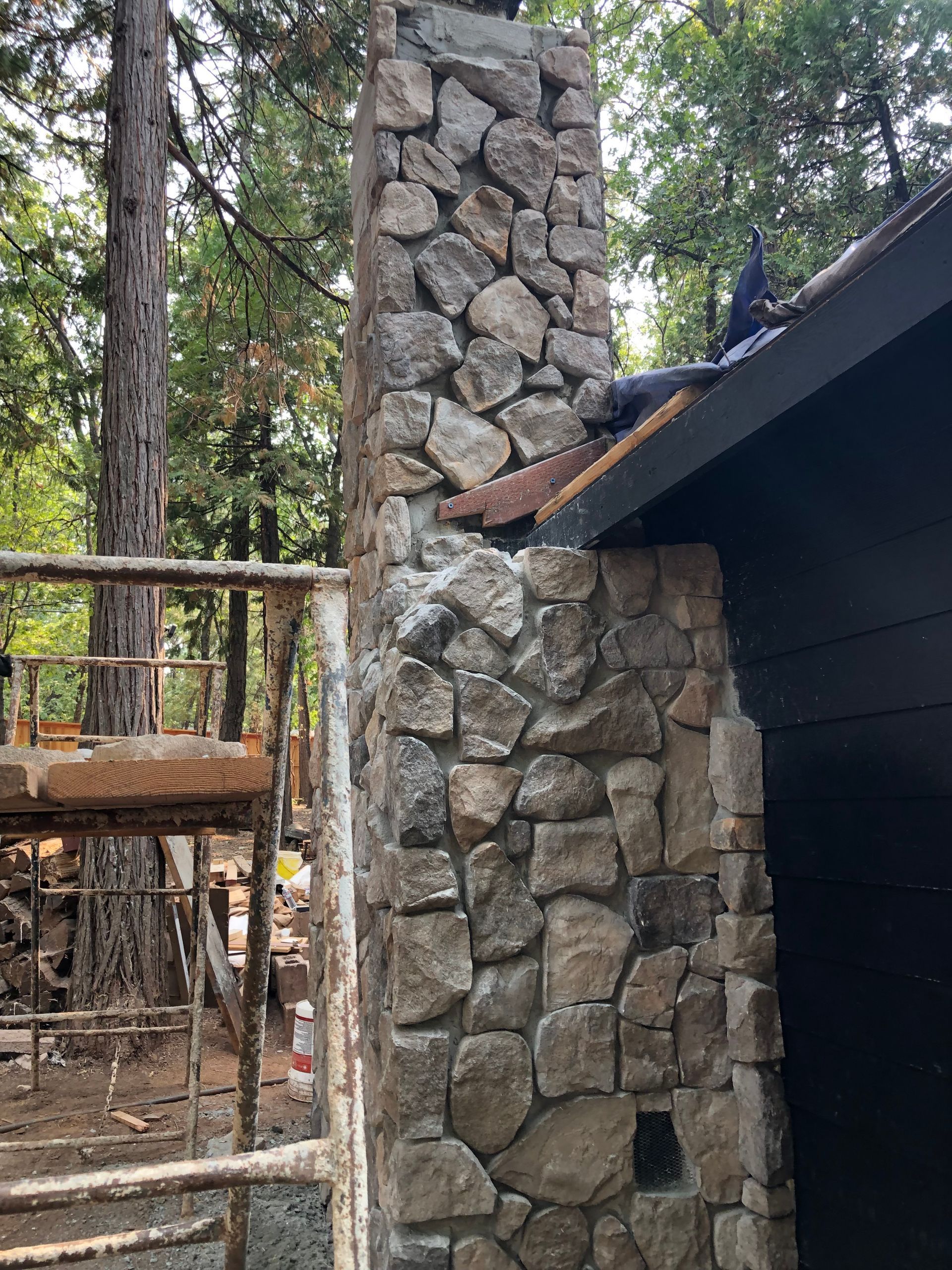 A stone chimney stands against a black wall next to construction scaffolding in a wooded area.