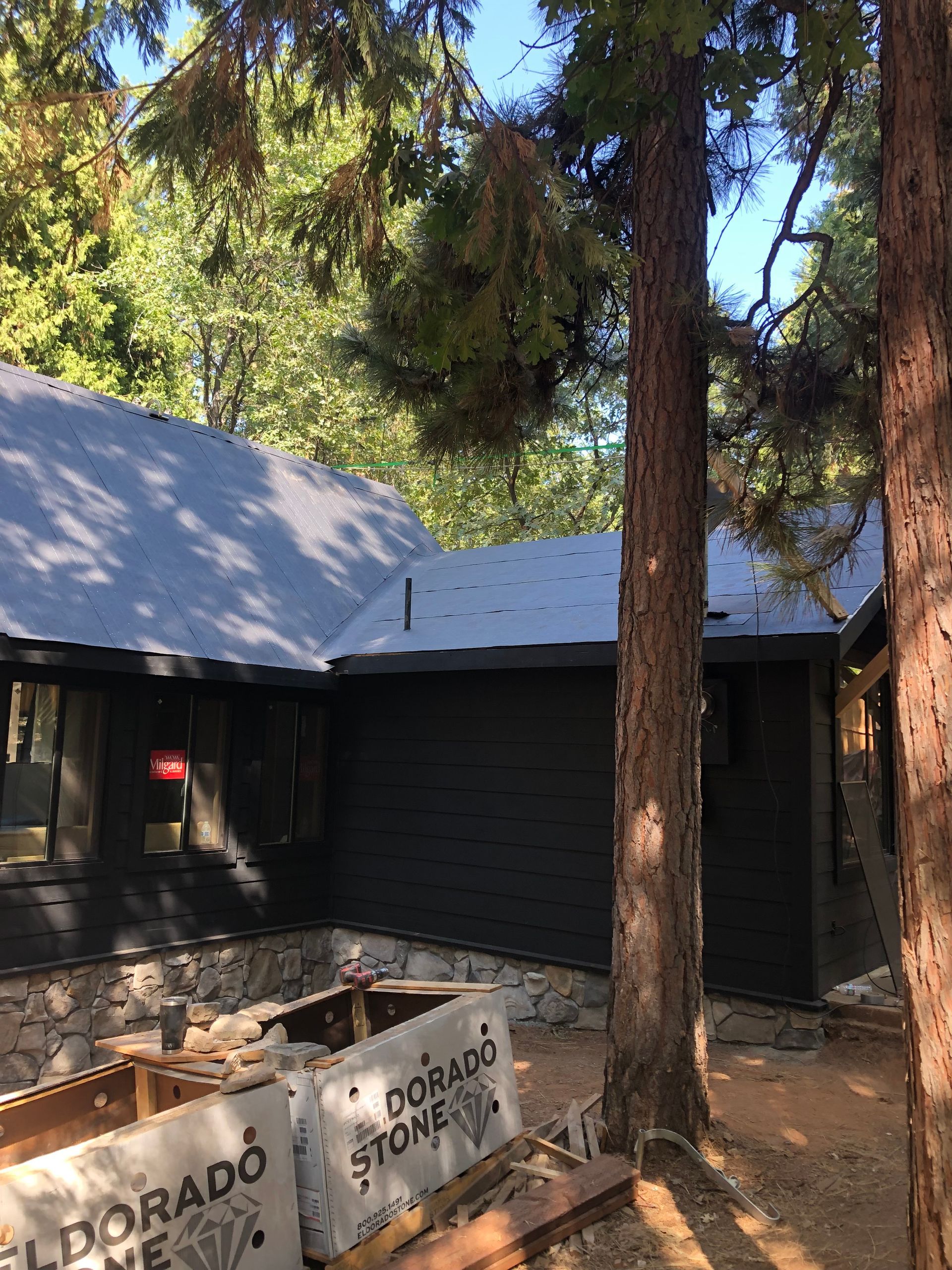A black-sided house under renovation with a new grey roof, surrounded by pine trees and Eldorado Stone supplies.