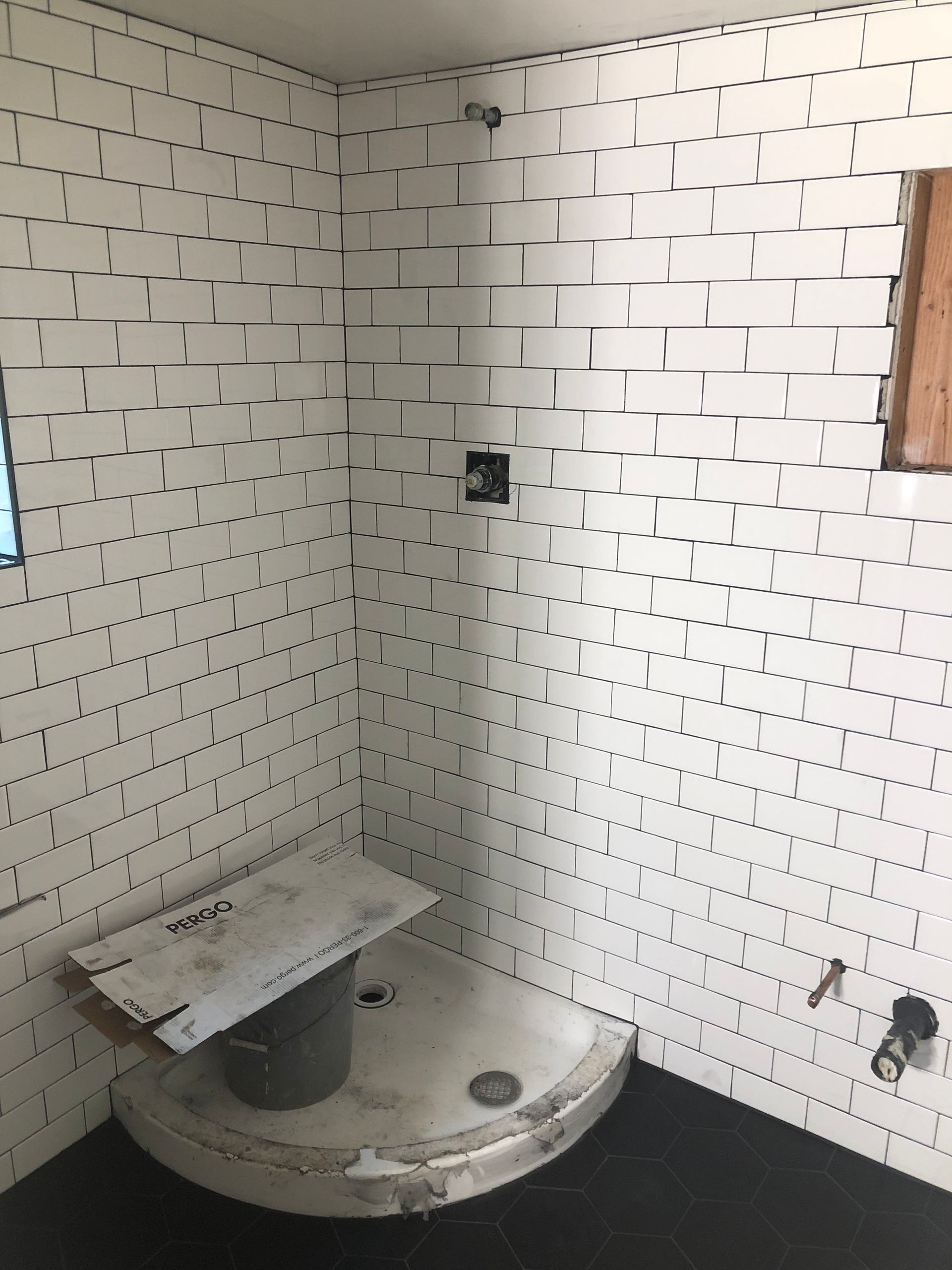 A partially renovated corner shower with white subway tiles, a round shower pan, and exposed plumbing against black floors.