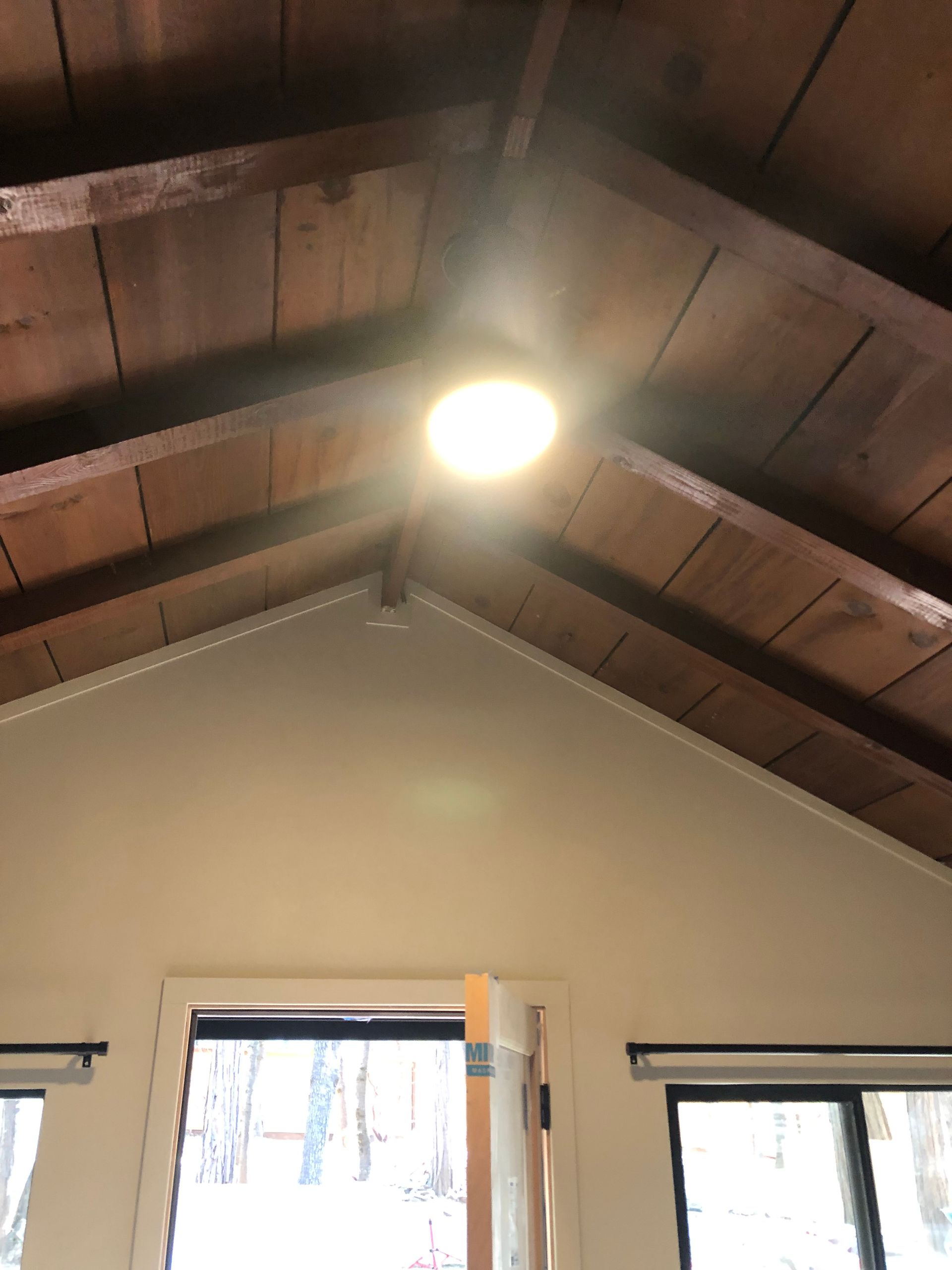 A low-angle view of a room with a peaked ceiling featuring exposed dark wooden beams, a bright overhead light, and windows.