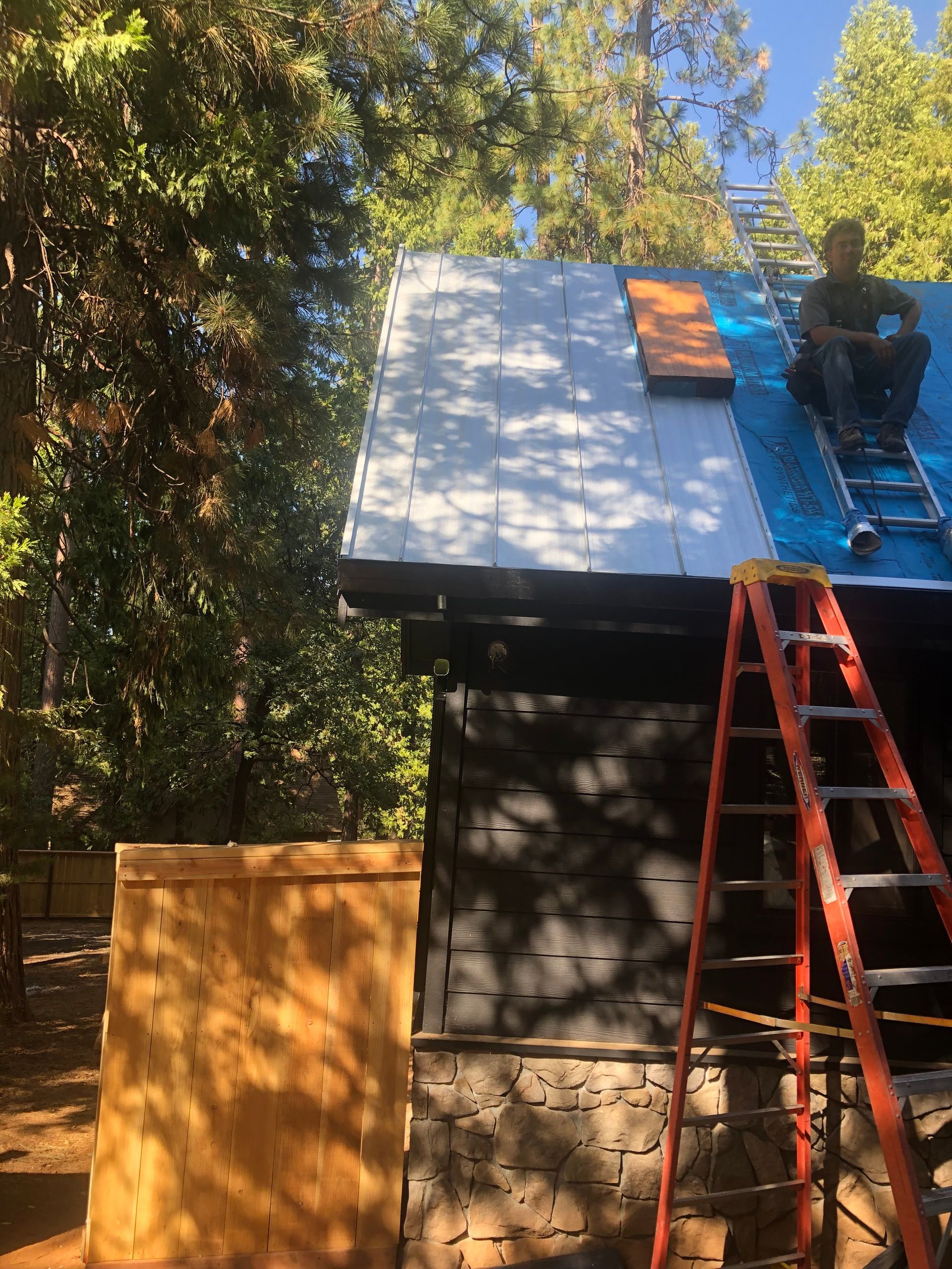 A person sits on a steep roof under construction, installing metal roofing panels beside a ladder near trees.