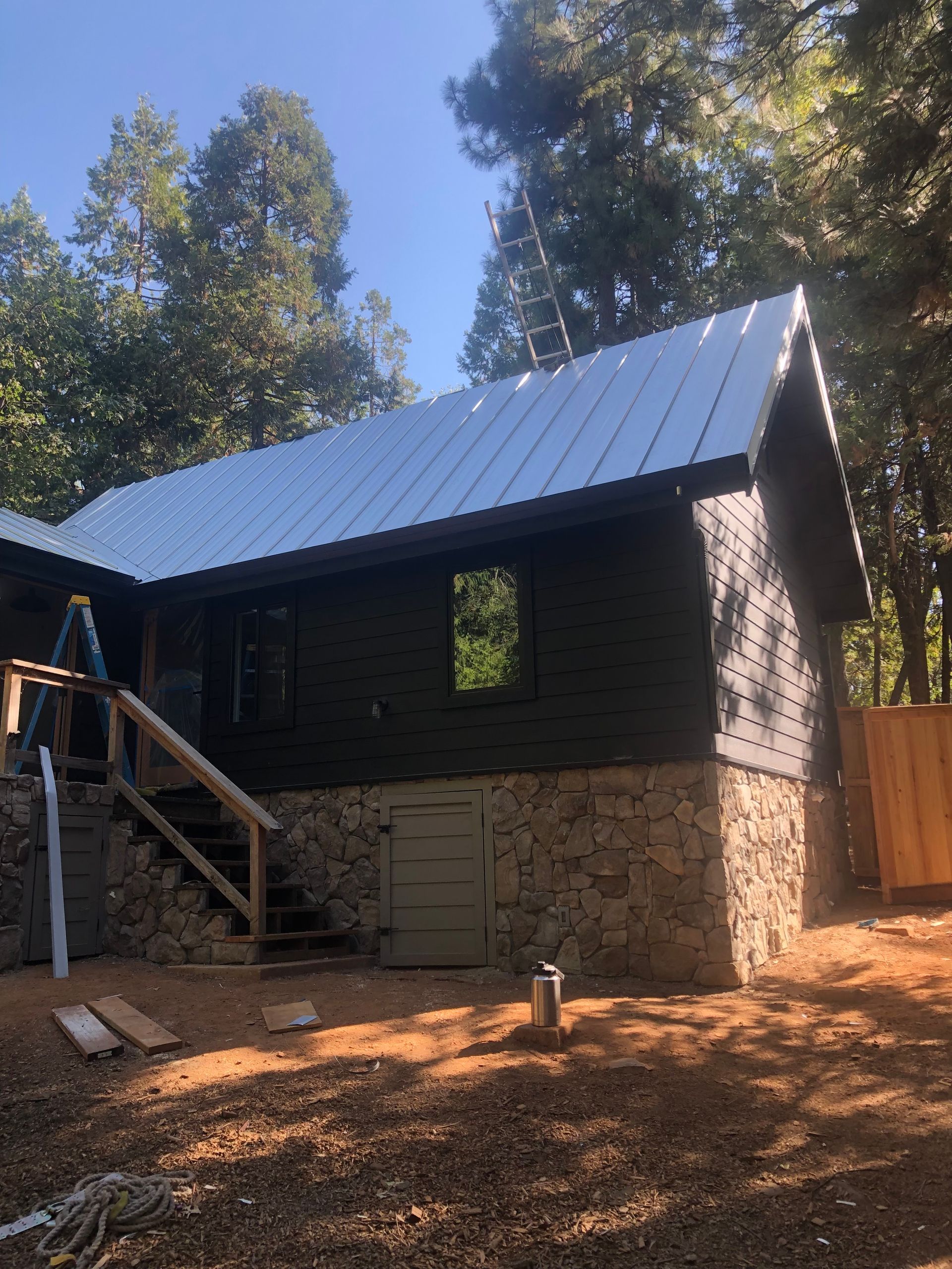 A dark-sided cabin with a metal roof and stone foundation sits in a wooded area with stairs leading to a side entrance.