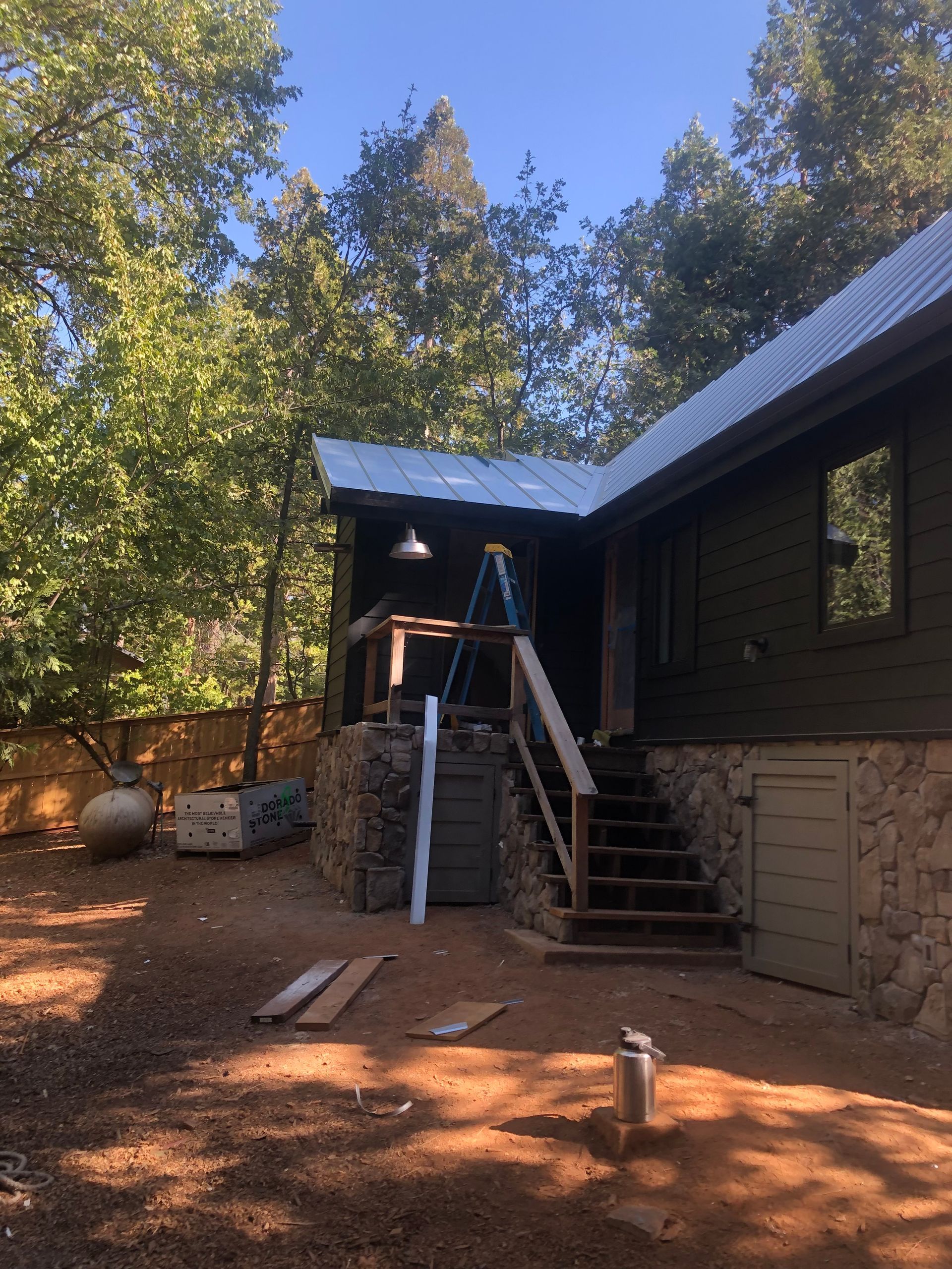 A dark brown cabin with a white metal roof and stone foundation, featuring an exterior wooden staircase and a ladder.