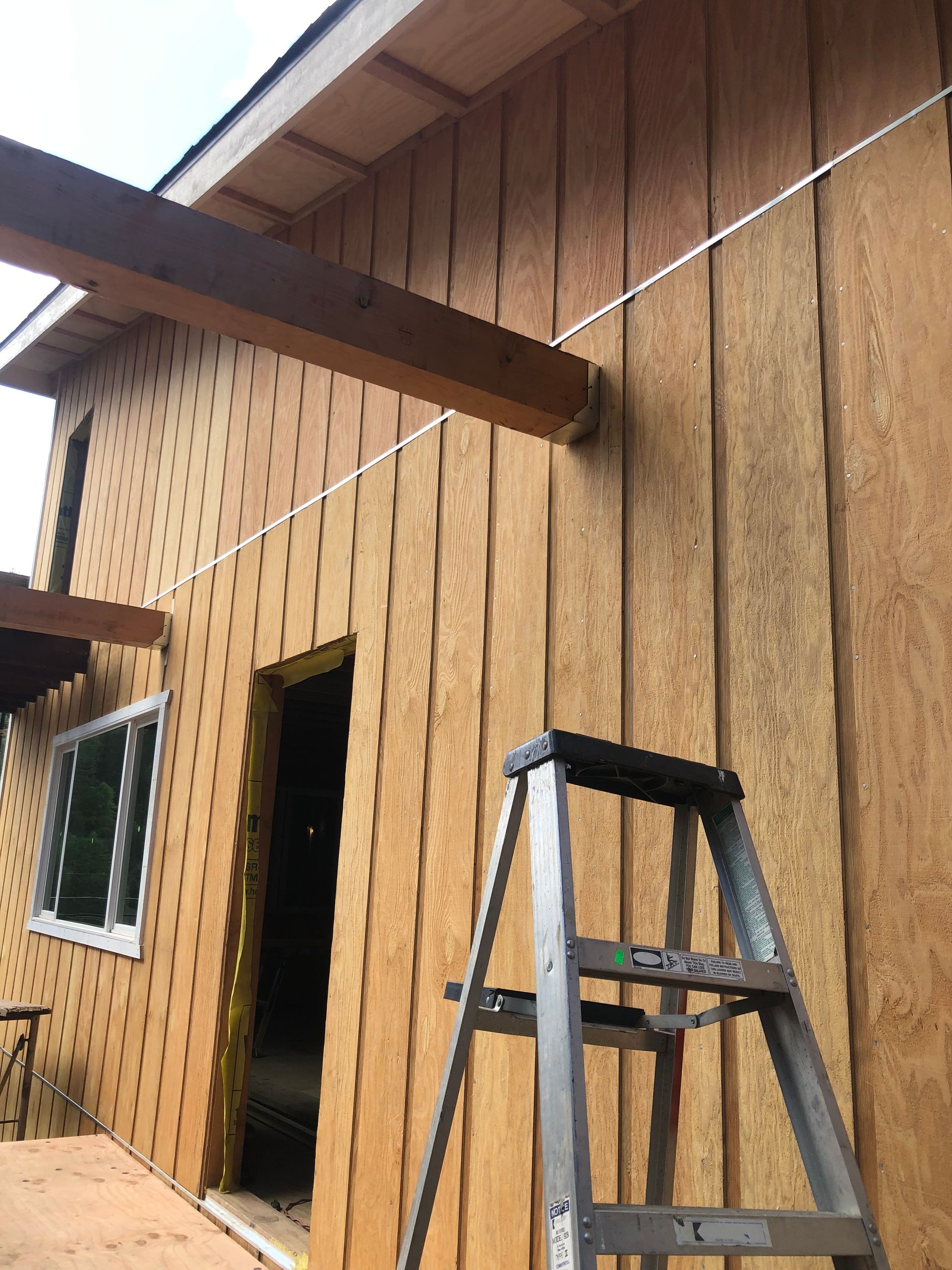 A stepladder sits outside a home under construction with unfinished wooden board-and-batten siding and an exposed beam.