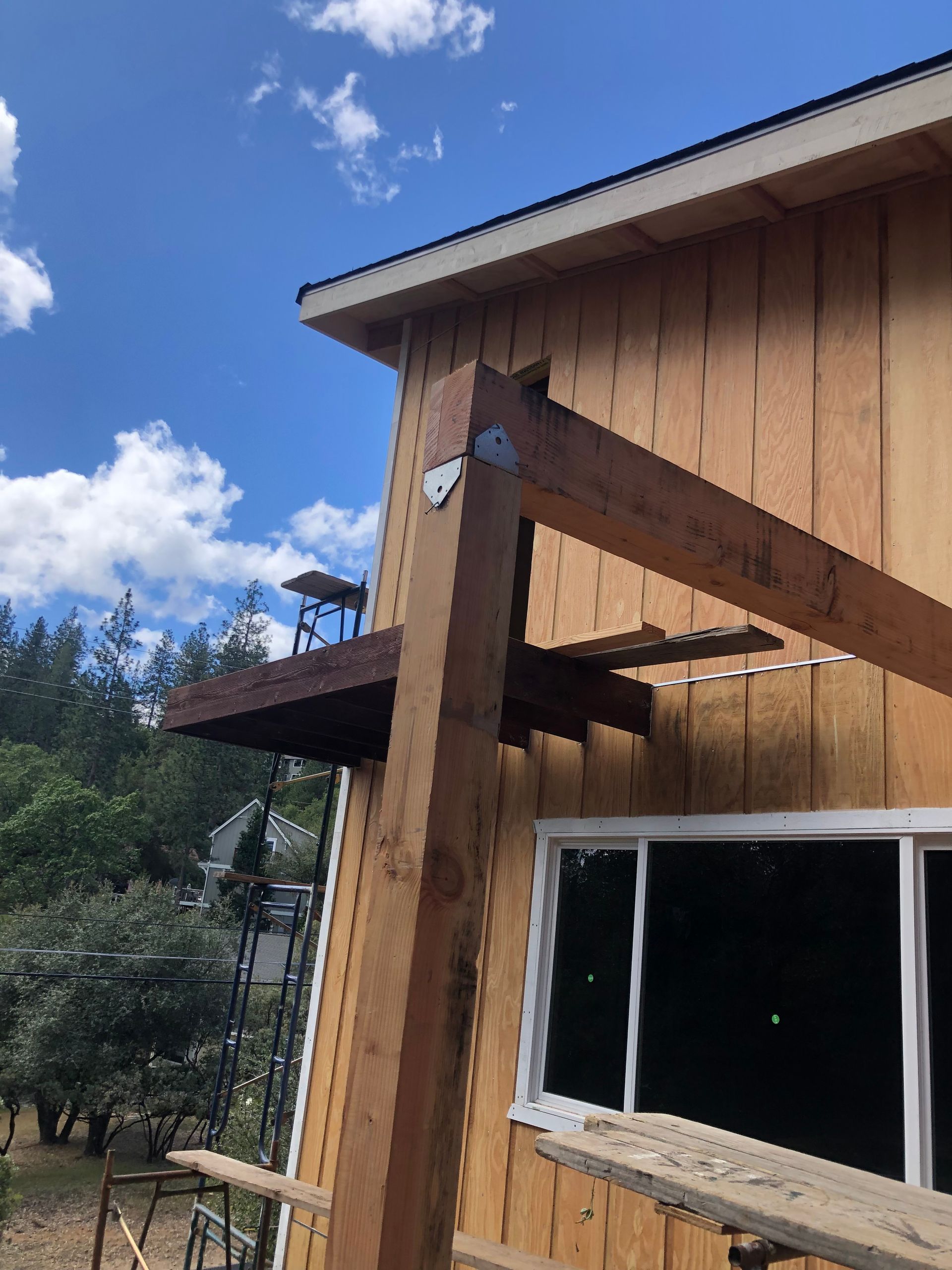 A vertical wooden post is connected to a horizontal beam with a metal bracket on the exterior of a wood-sided building.