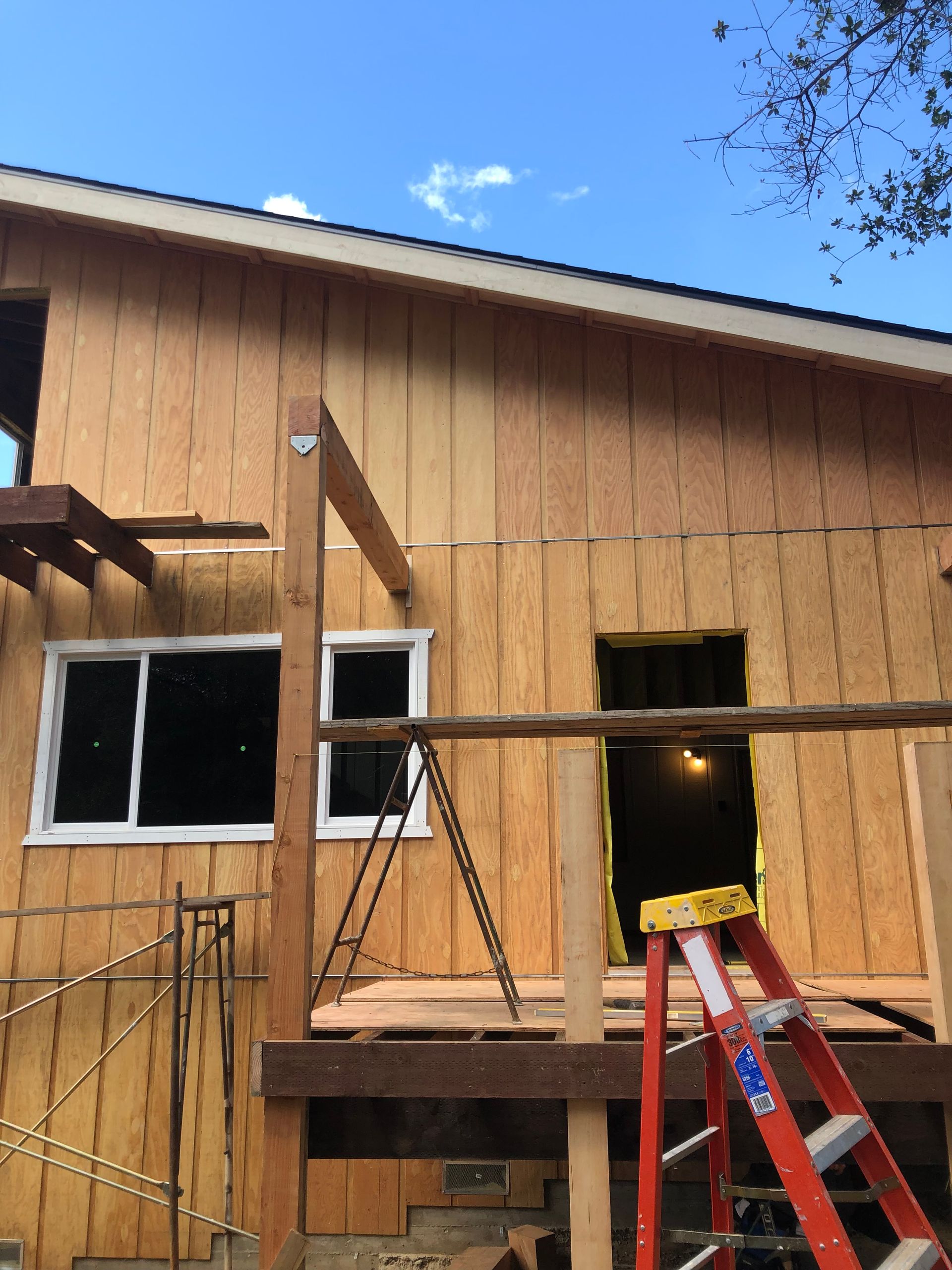 Exterior view of a residential building under construction with exposed plywood siding, a new window, and a red ladder.