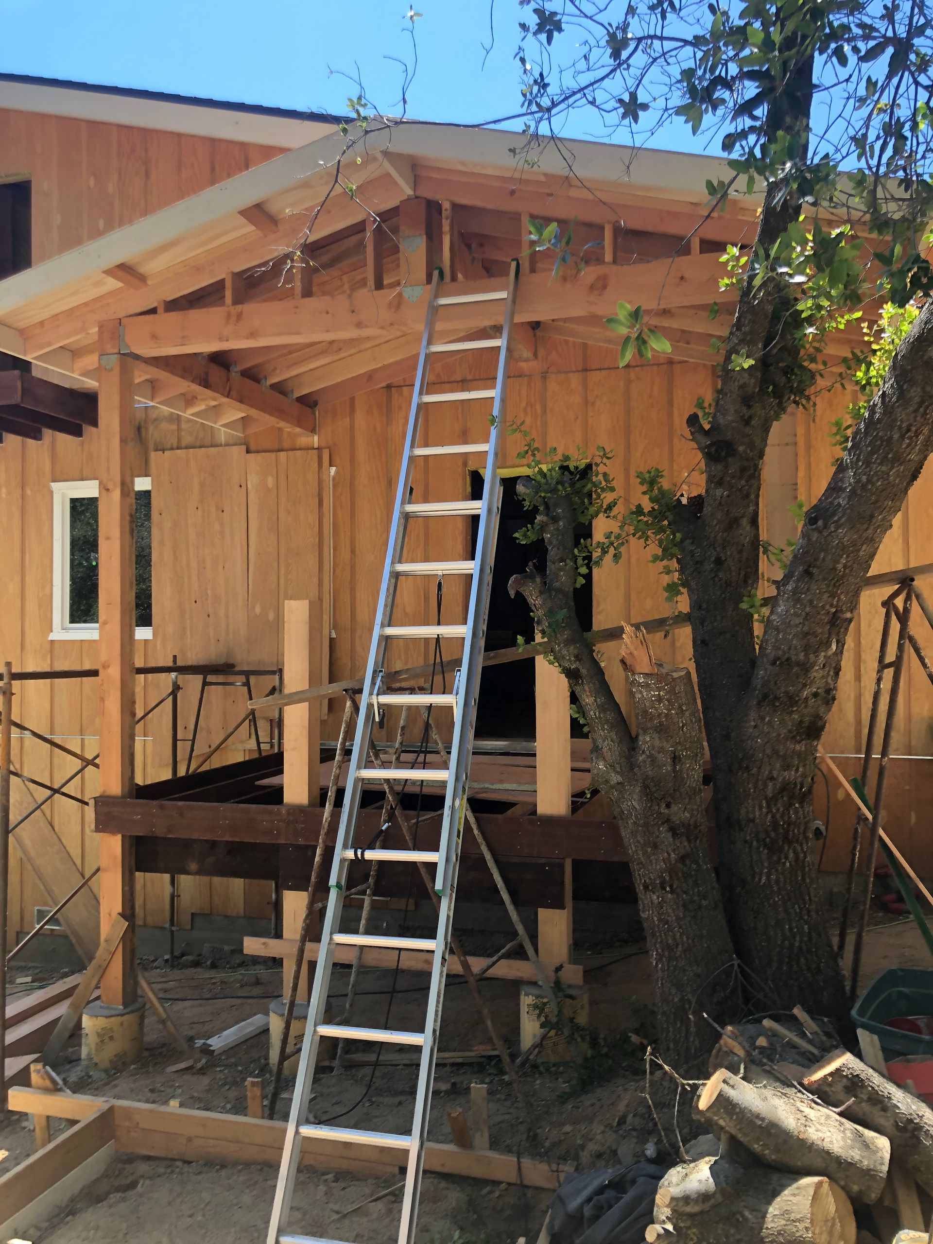 A wooden house structure under construction, featuring an unfinished deck, framing, and a tall metal ladder leaning on it.