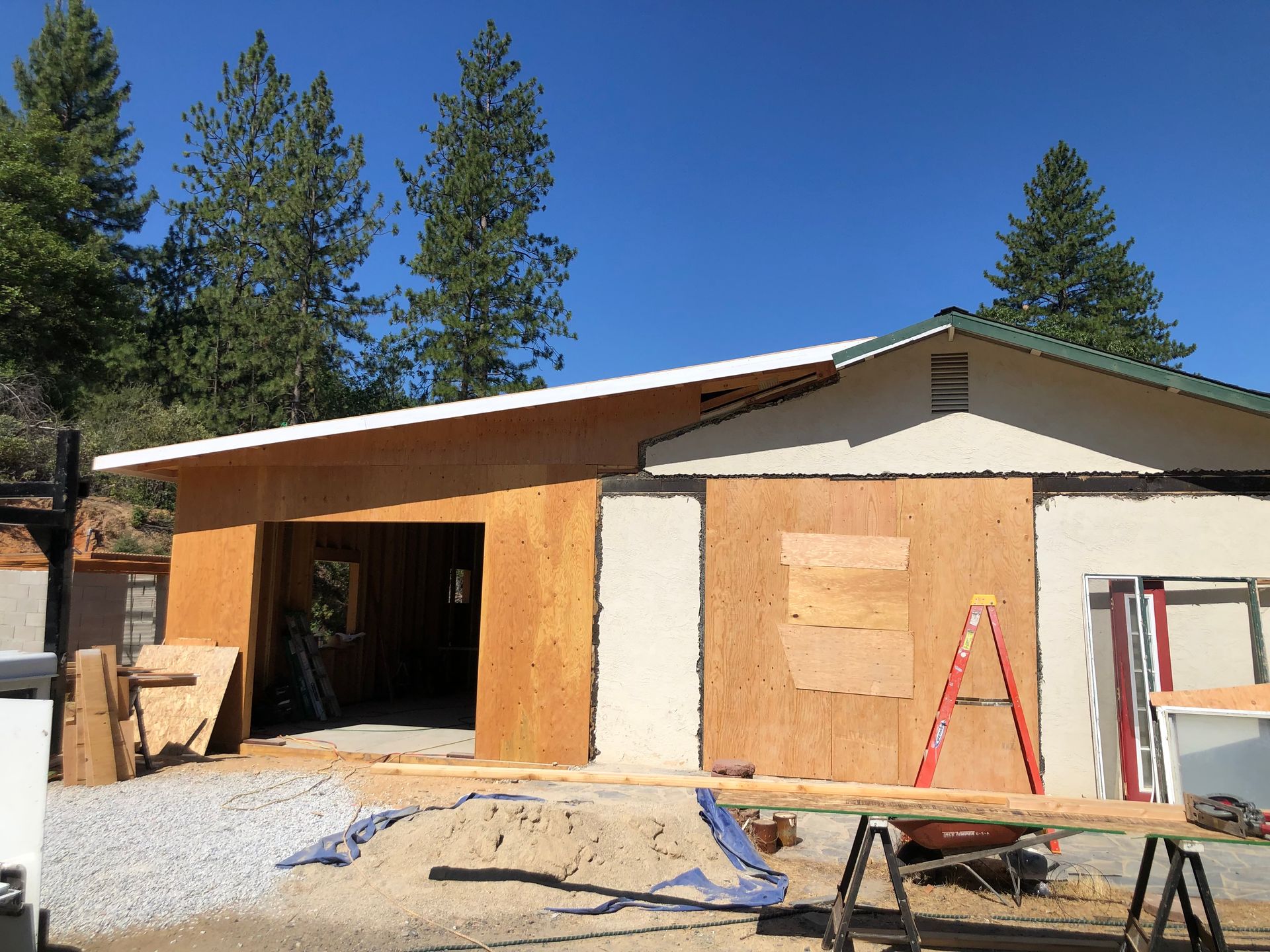Exterior view of a home under construction with plywood wall panels, an open garage bay, and a red ladder on site.