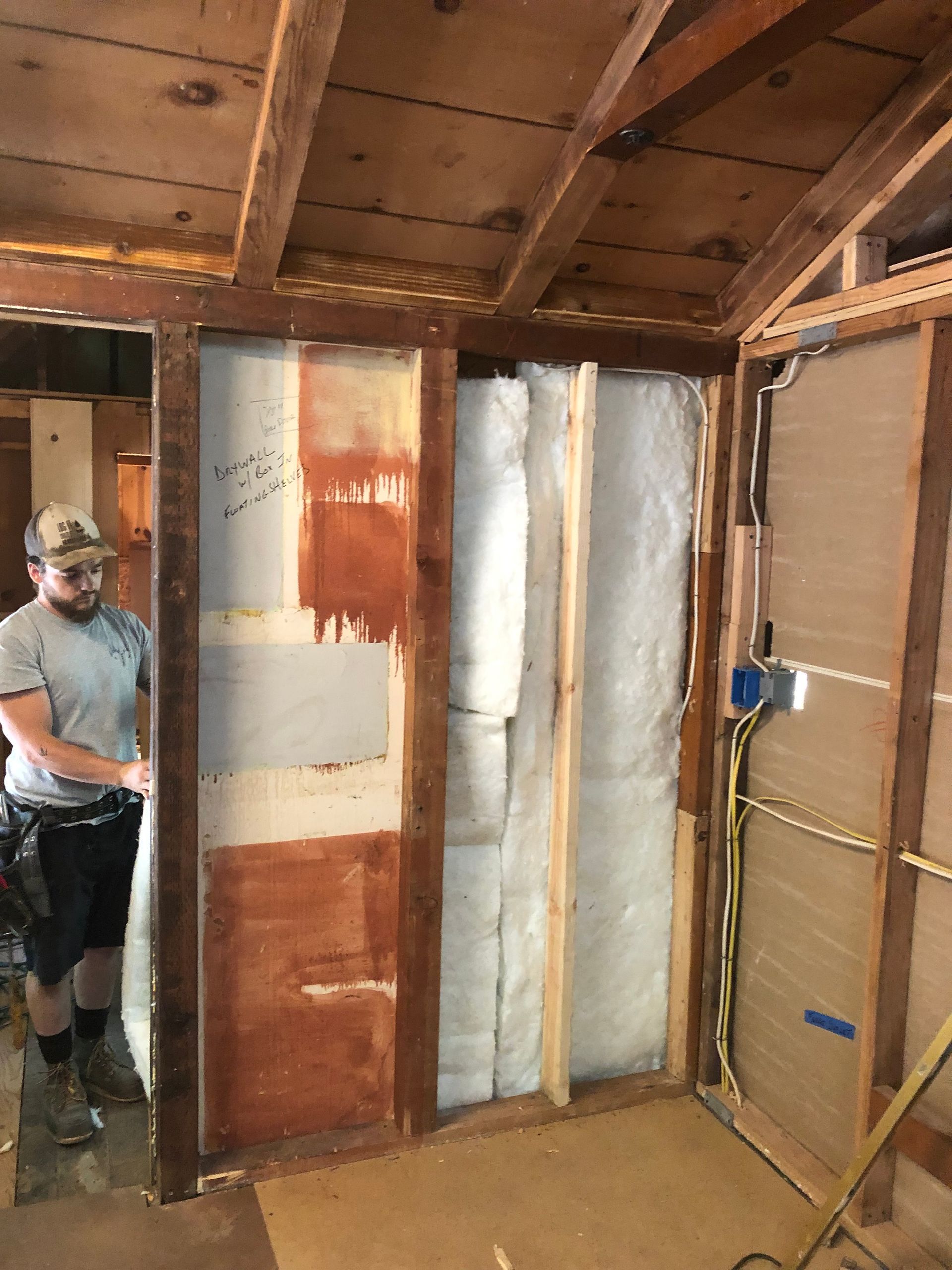 A person stands in a construction site interior with wood wall framing, exposed fiberglass insulation, and electrical wires.