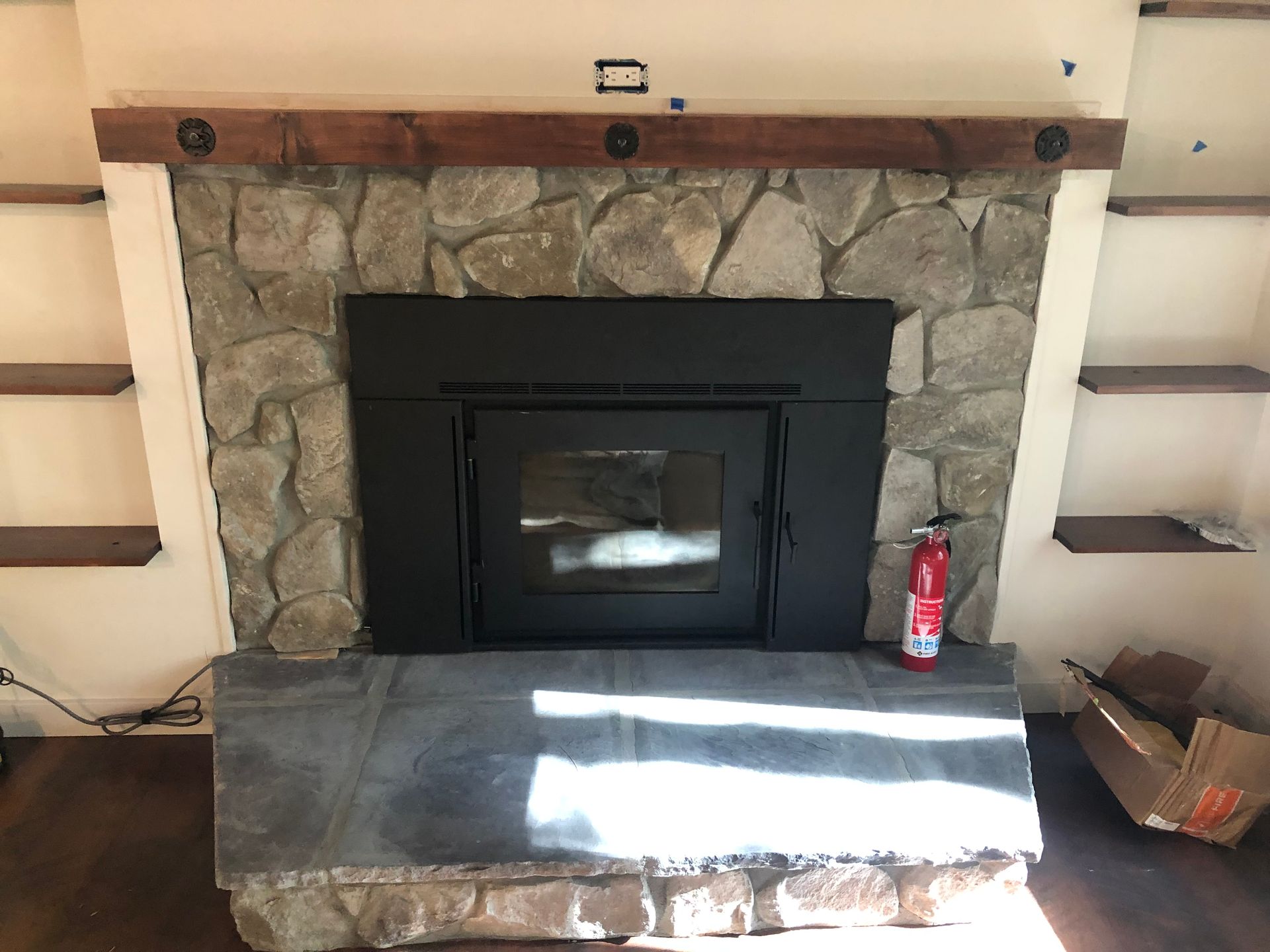 A black wood-burning fireplace insert with a stone surround, slate hearth, and wooden mantel, flanked by wall shelves.