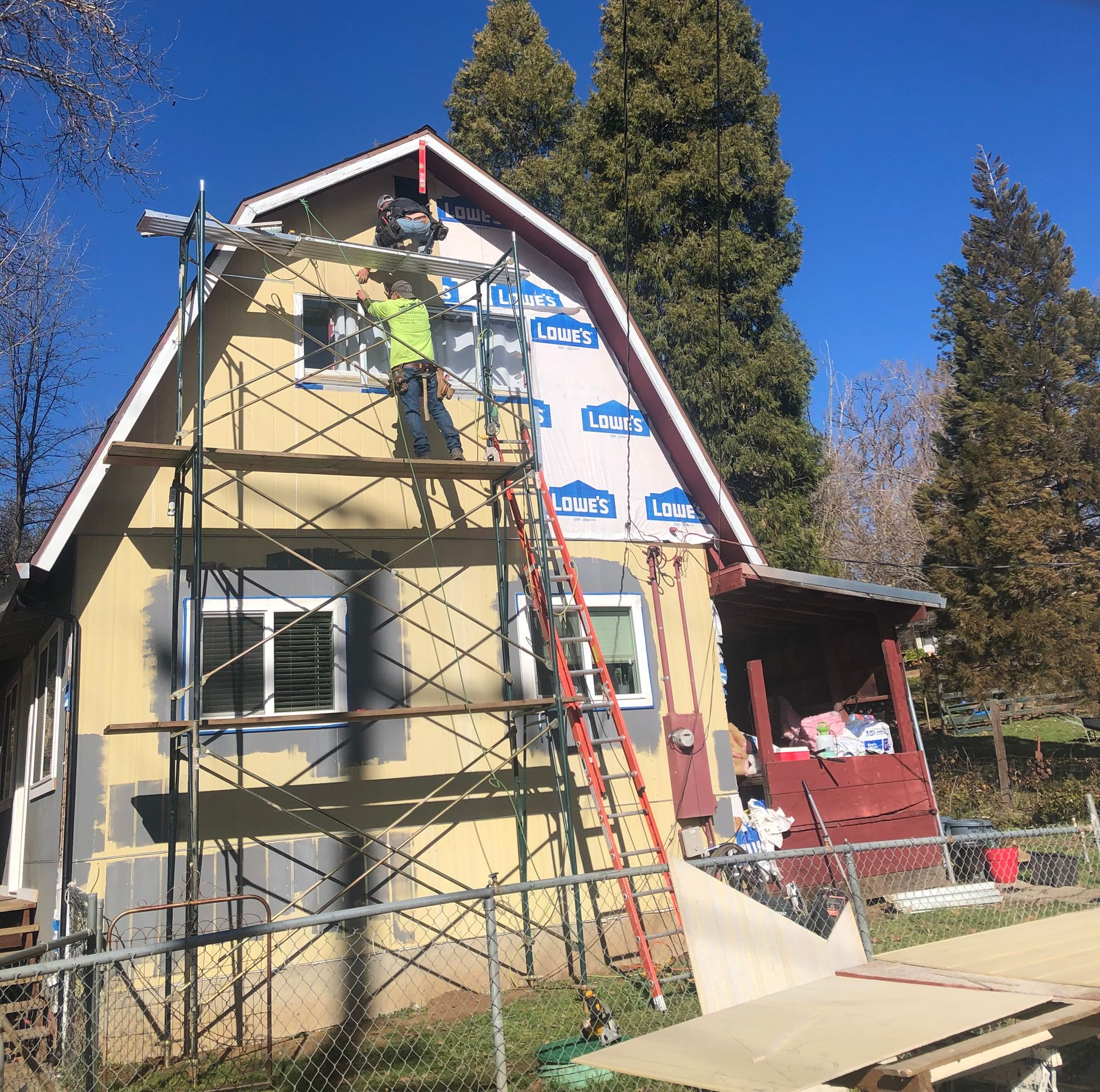 A worker in a high-visibility vest stands on scaffolding while installing sheathing on the gable of a house.