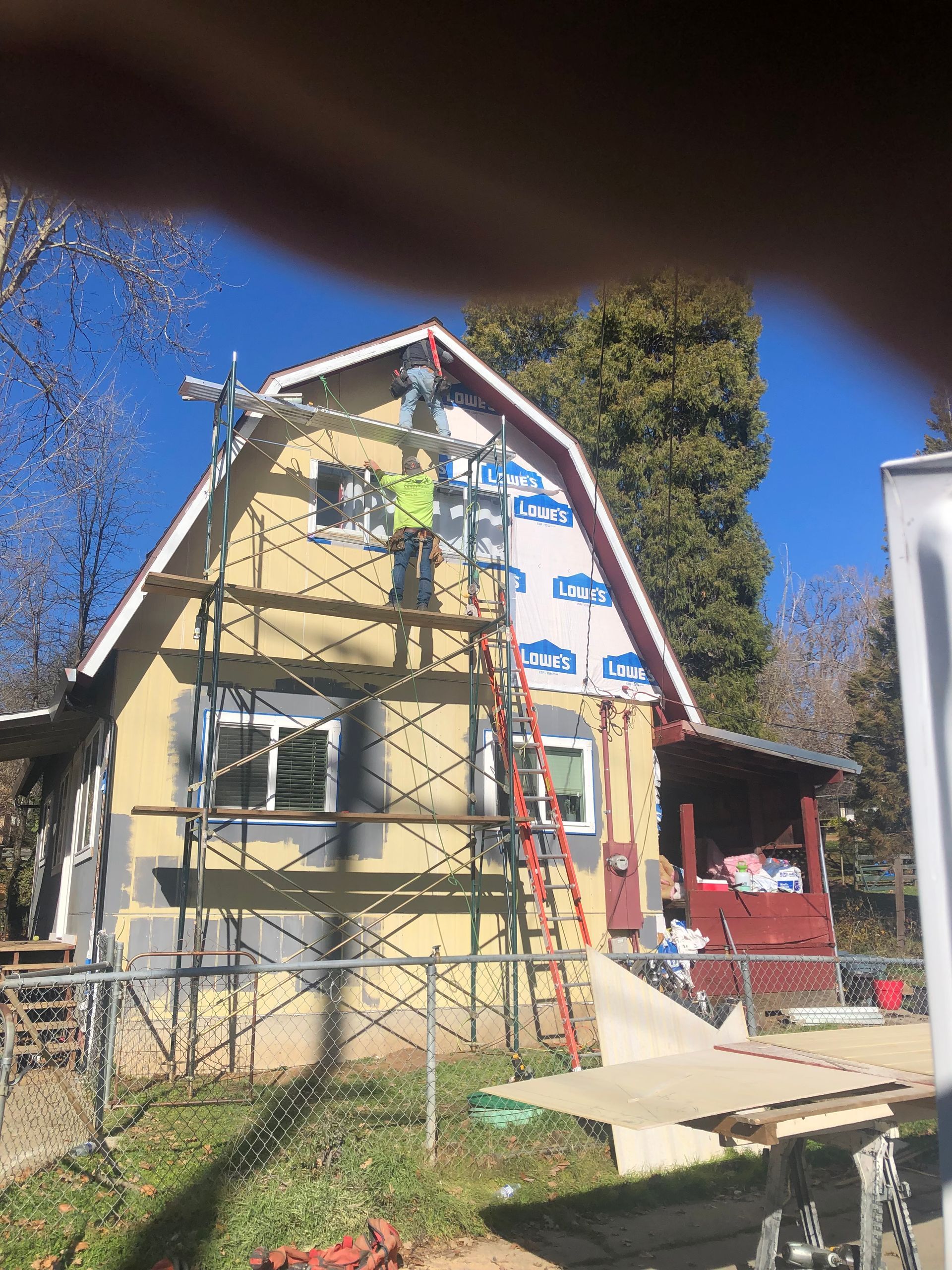 A worker in a yellow vest stands on scaffolding while repairing the upper exterior of a house under a clear blue sky.
