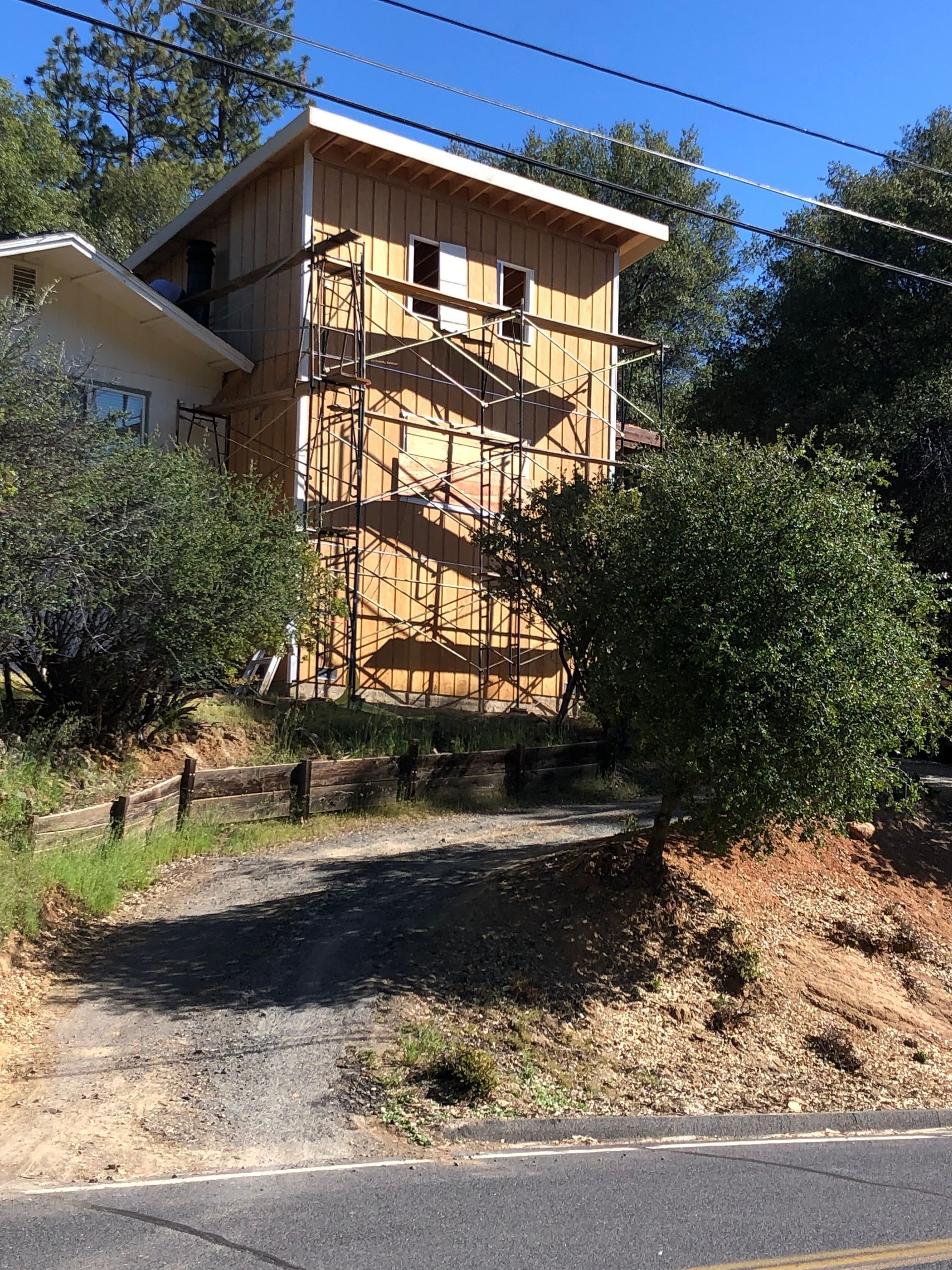 A two-story house under construction with scaffolding against a light wood-sided wall, situated on a sloped, tree-lined lot.