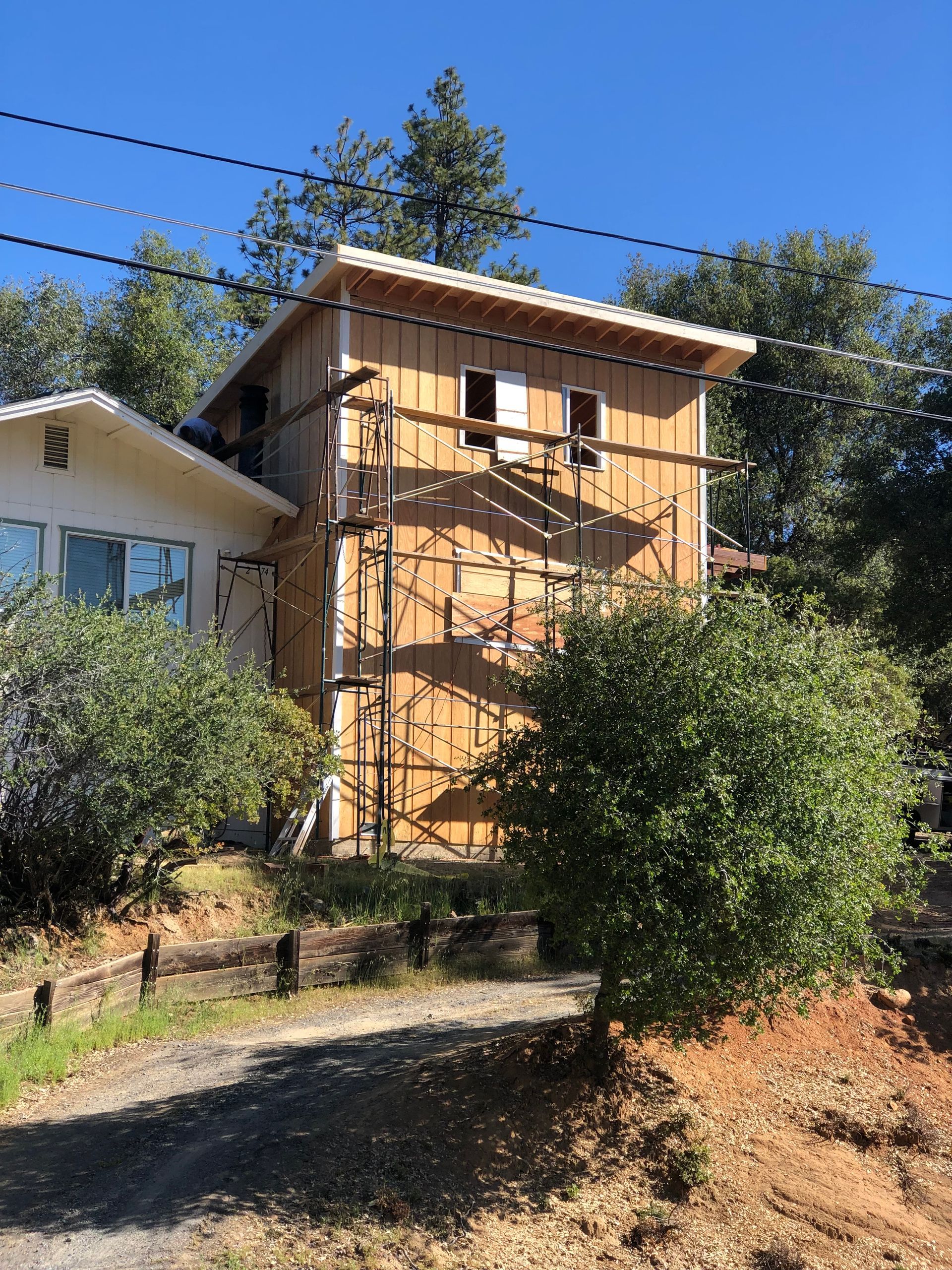 A multi-story home under construction with wooden siding and scaffolding in a sunny, wooded outdoor setting.