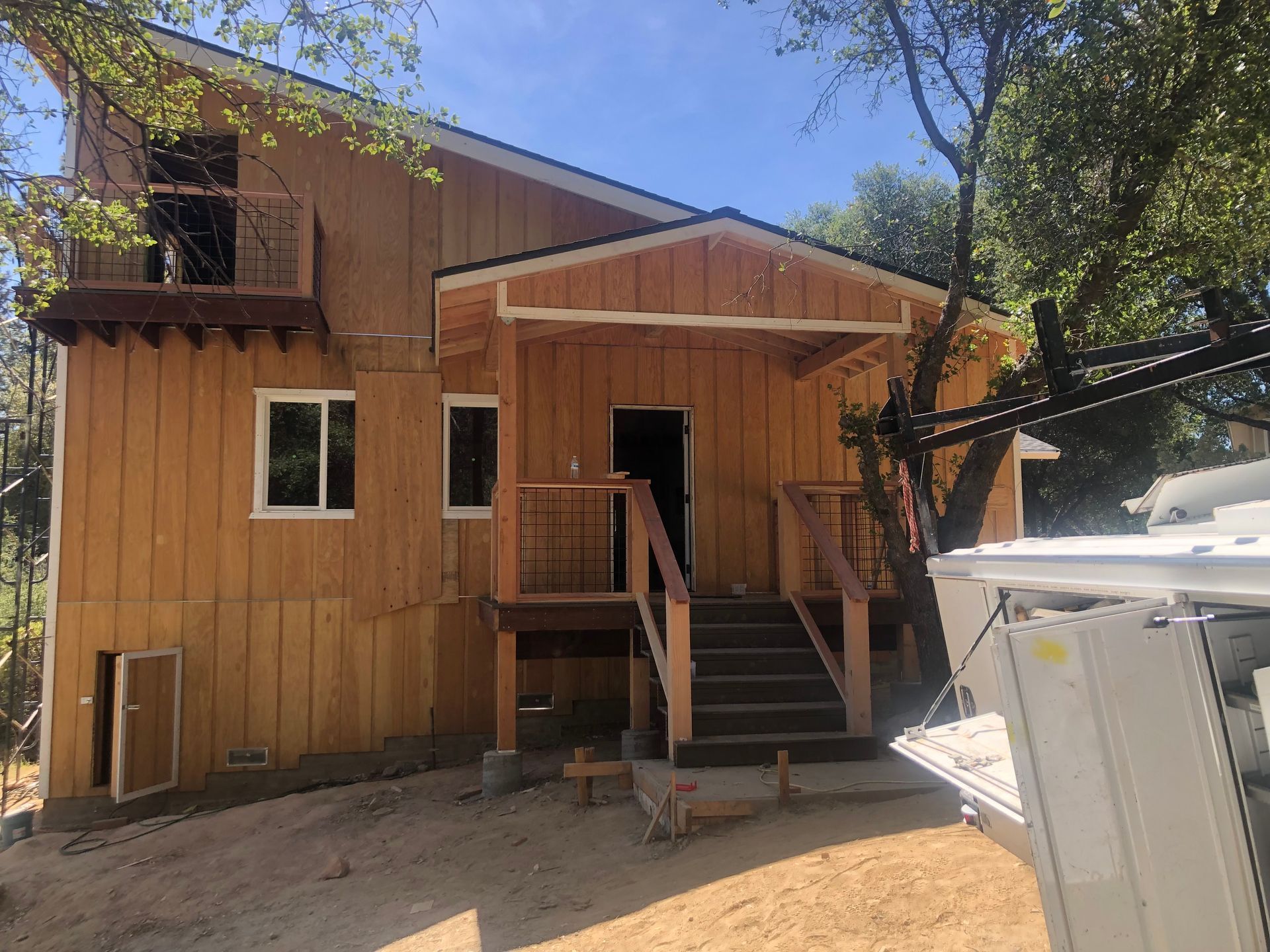 A two-story wood-sided house with a front porch, stairs, and a balcony under construction in a wooded area.