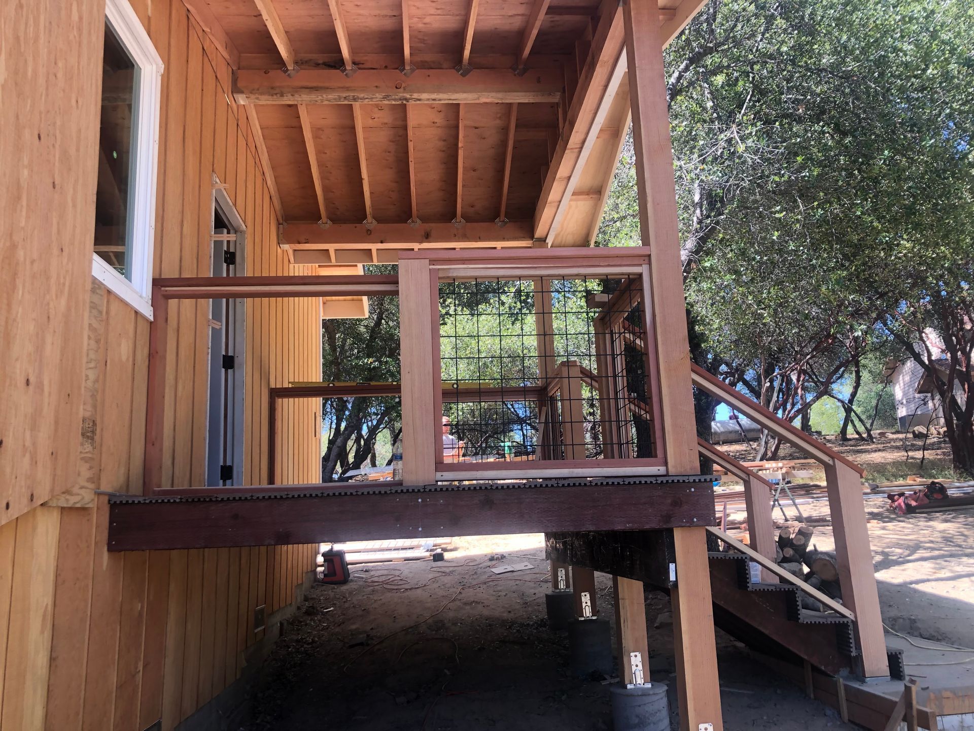 A wooden house deck under construction with a frame railing and stairs leading down to a dirt yard with trees.