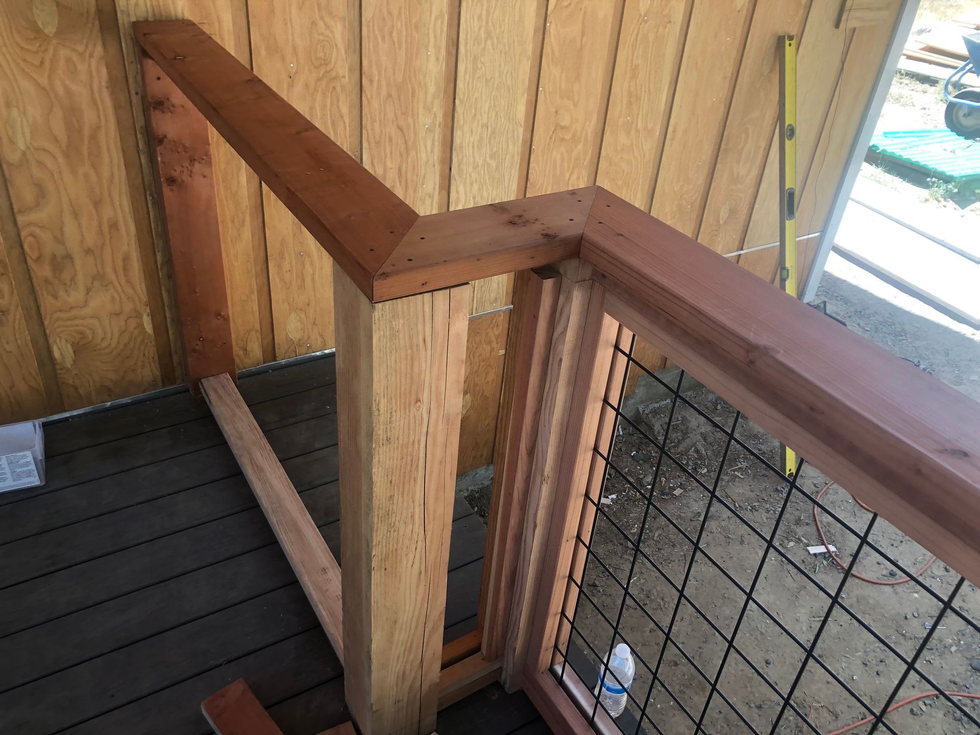A wooden deck railing with a corner miter joint, wire mesh panels, and a vertical support post against a wooden wall.