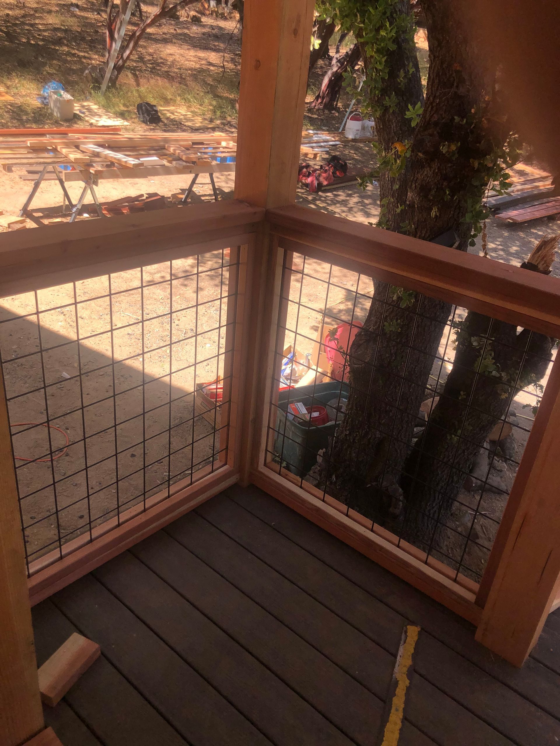A wooden deck corner featuring railings with a metal grid panel and a large tree trunk visible in the background.