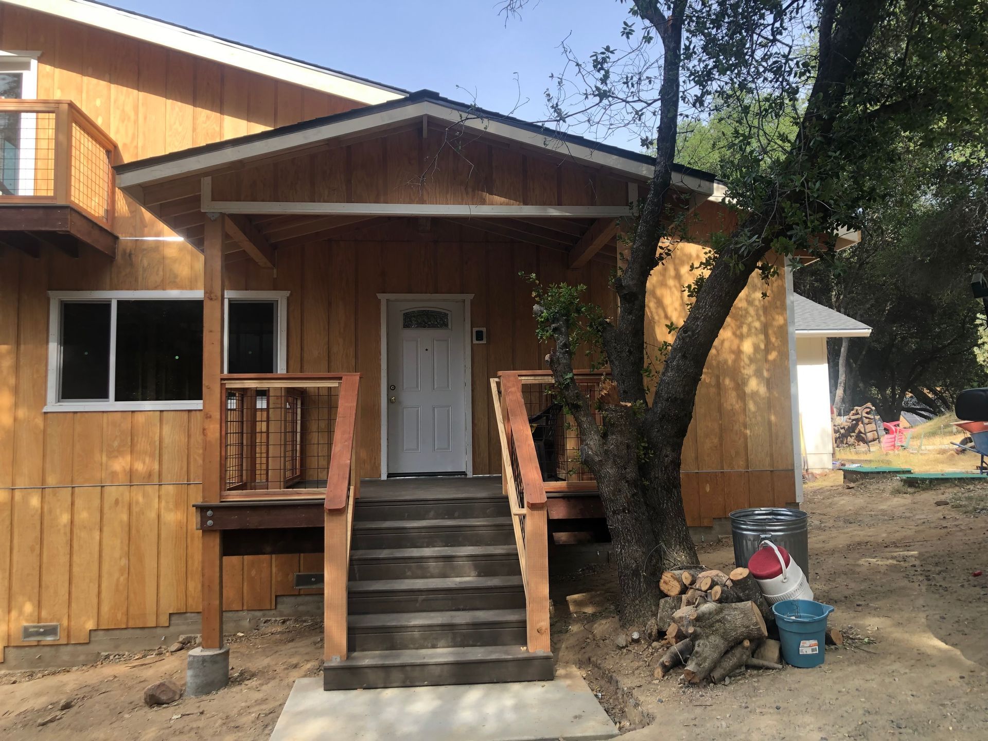 Wooden house exterior featuring a front porch with dark wooden stairs, a white front door, and a tree nearby.
