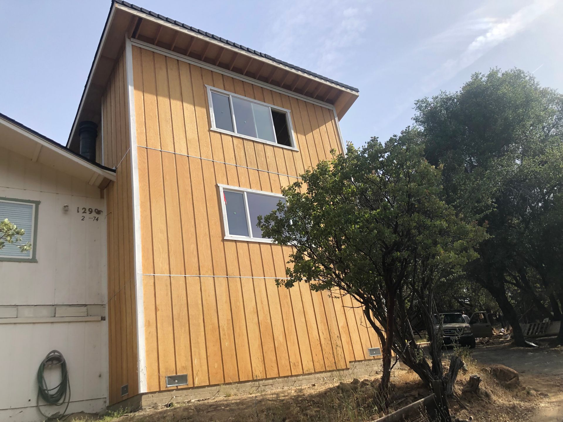 A two-story building exterior with vertical light-wood paneling, white trim, and two windows, set next to a tree.