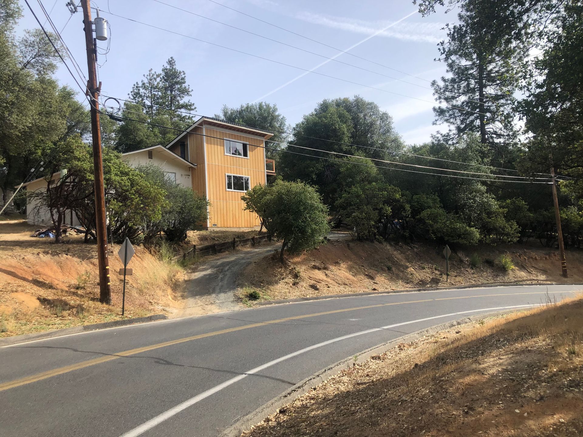 A two-story tan house sits set back on a hillside beside a paved two-lane road, surrounded by trees and dry vegetation.