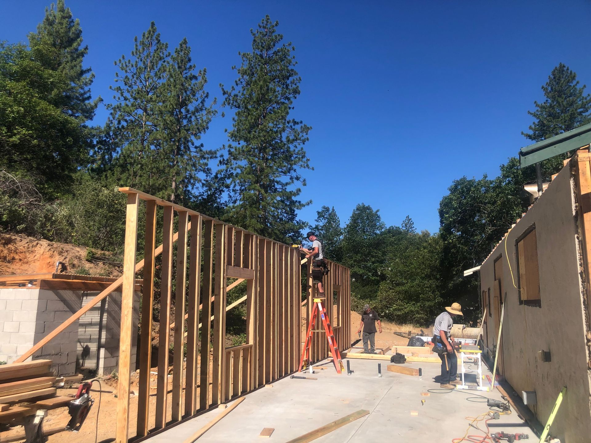 Workers construct a wooden frame for a building on a sunny, outdoor construction site near trees.