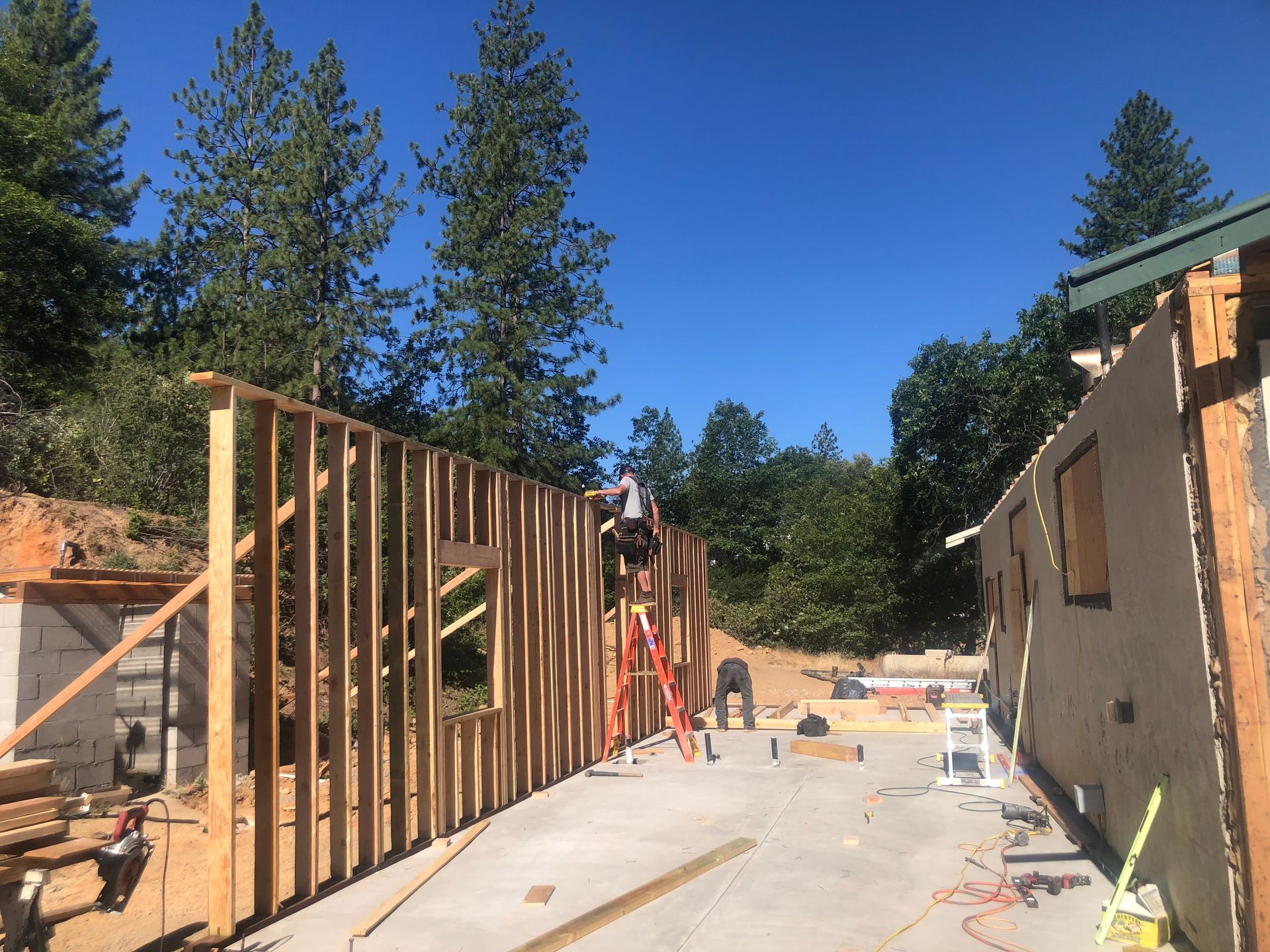 Construction site with a new wooden wall frame, workers, and tools on a sunny day near a forested hill.