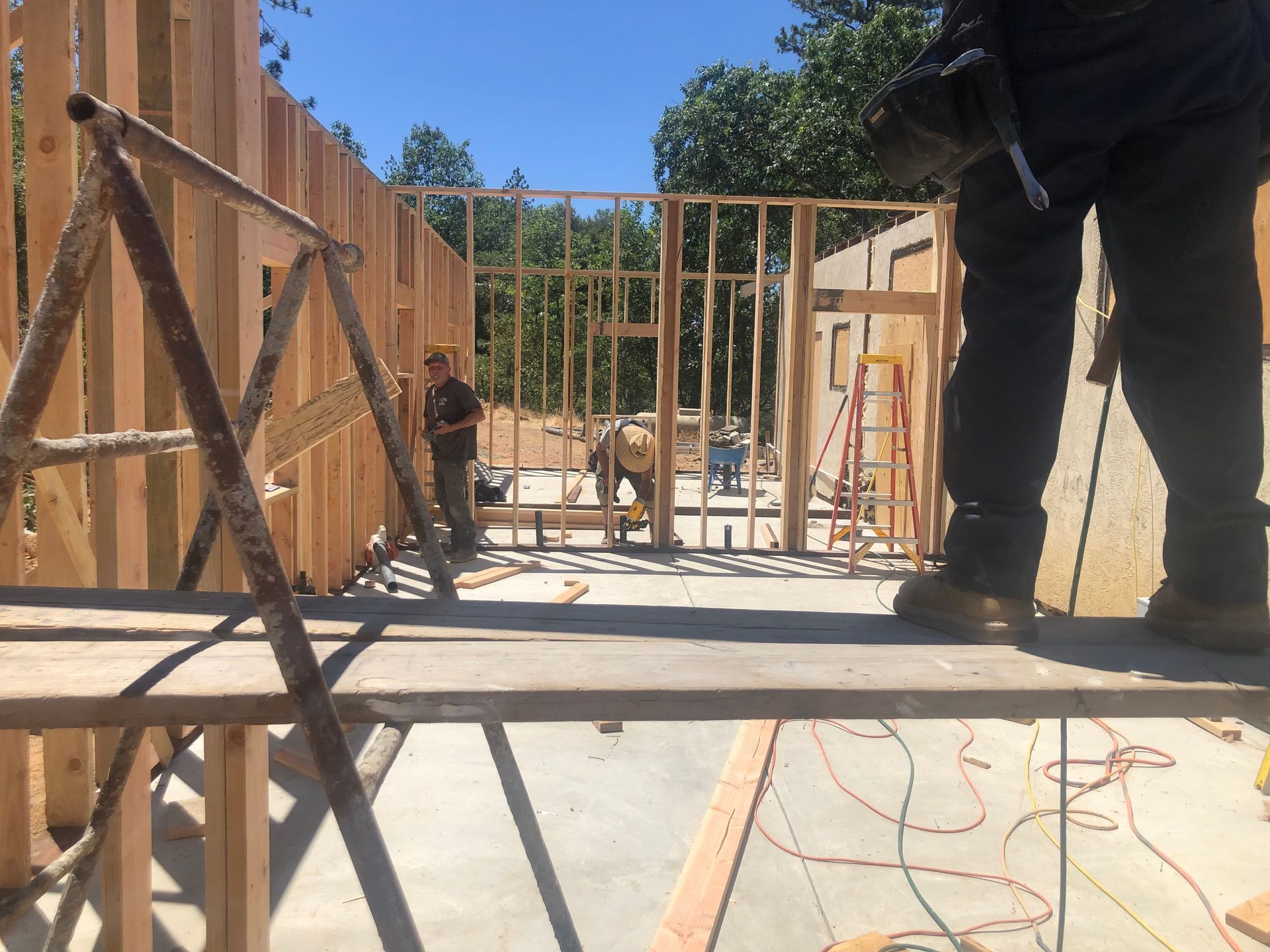 Construction workers frame a house structure outdoors on a sunny day, viewed from a wooden scaffolding platform.