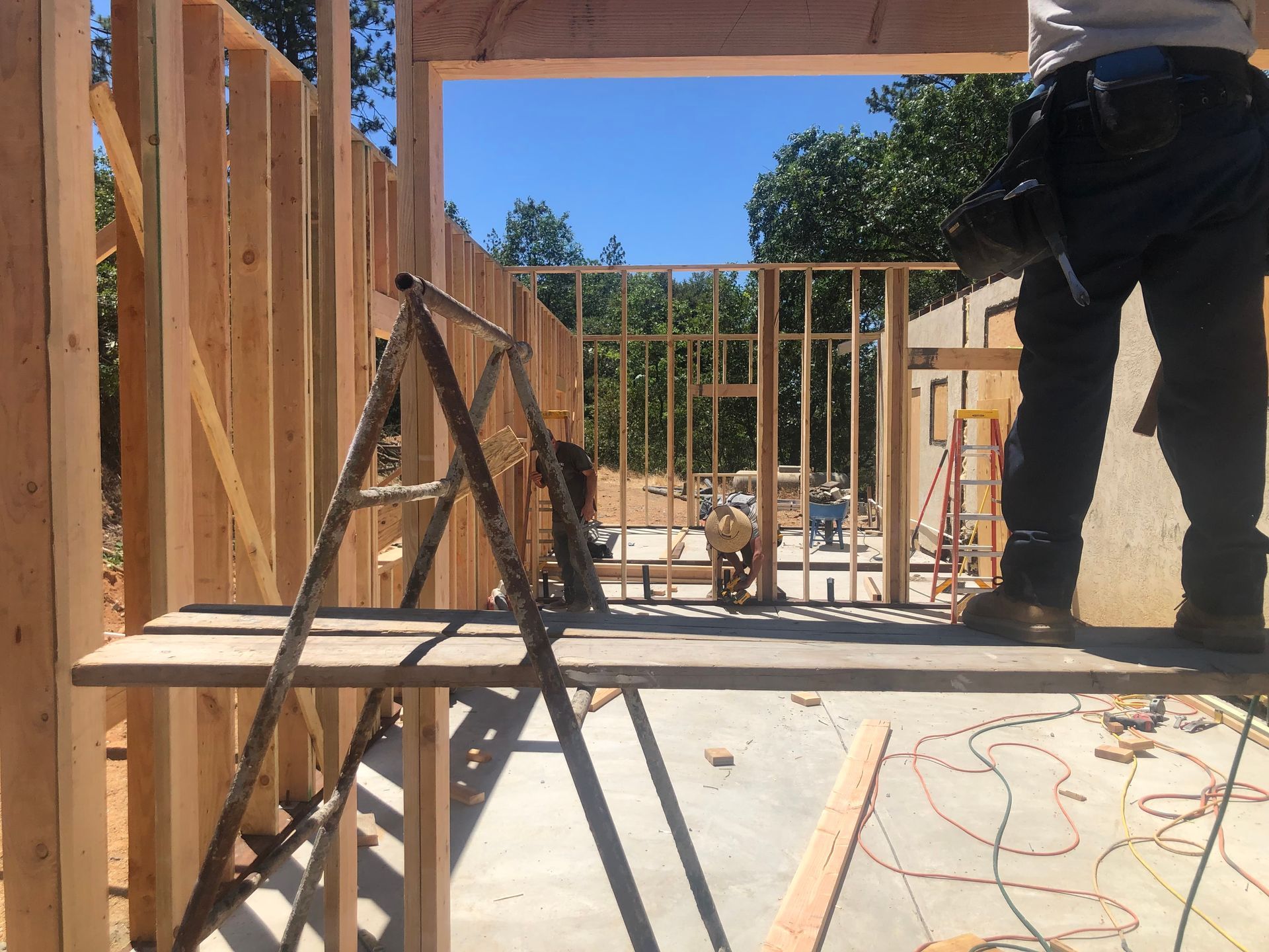A person stands on a wooden scaffolding plank, framing the wooden wall structure of a house under construction.