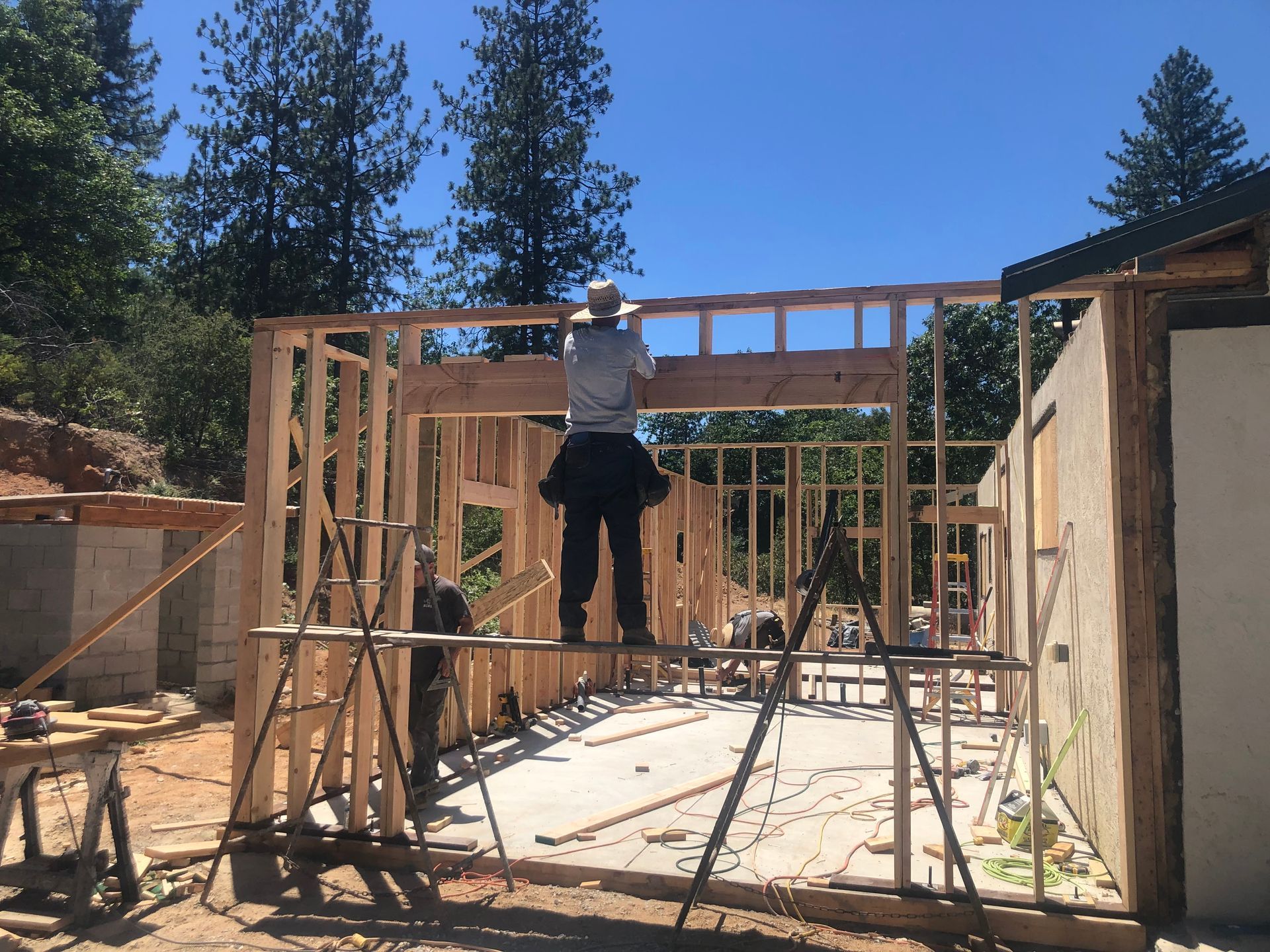 A worker standing on scaffolding installs a large wooden header beam onto the framed wall of a building under construction.