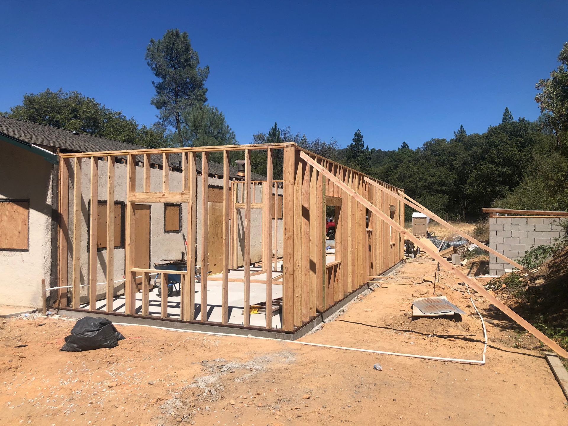 Wooden framing for a home addition stands on a construction site with a clear blue sky in the background.