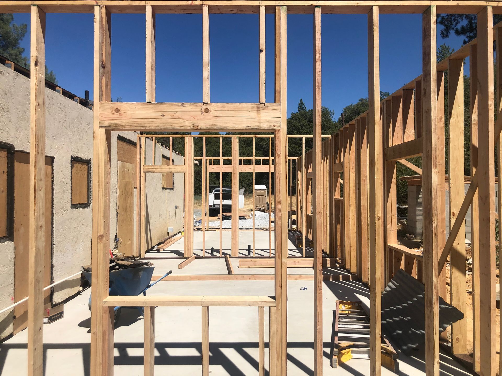 A view through wooden wall framing at a construction site, looking toward an open structure on a concrete foundation.