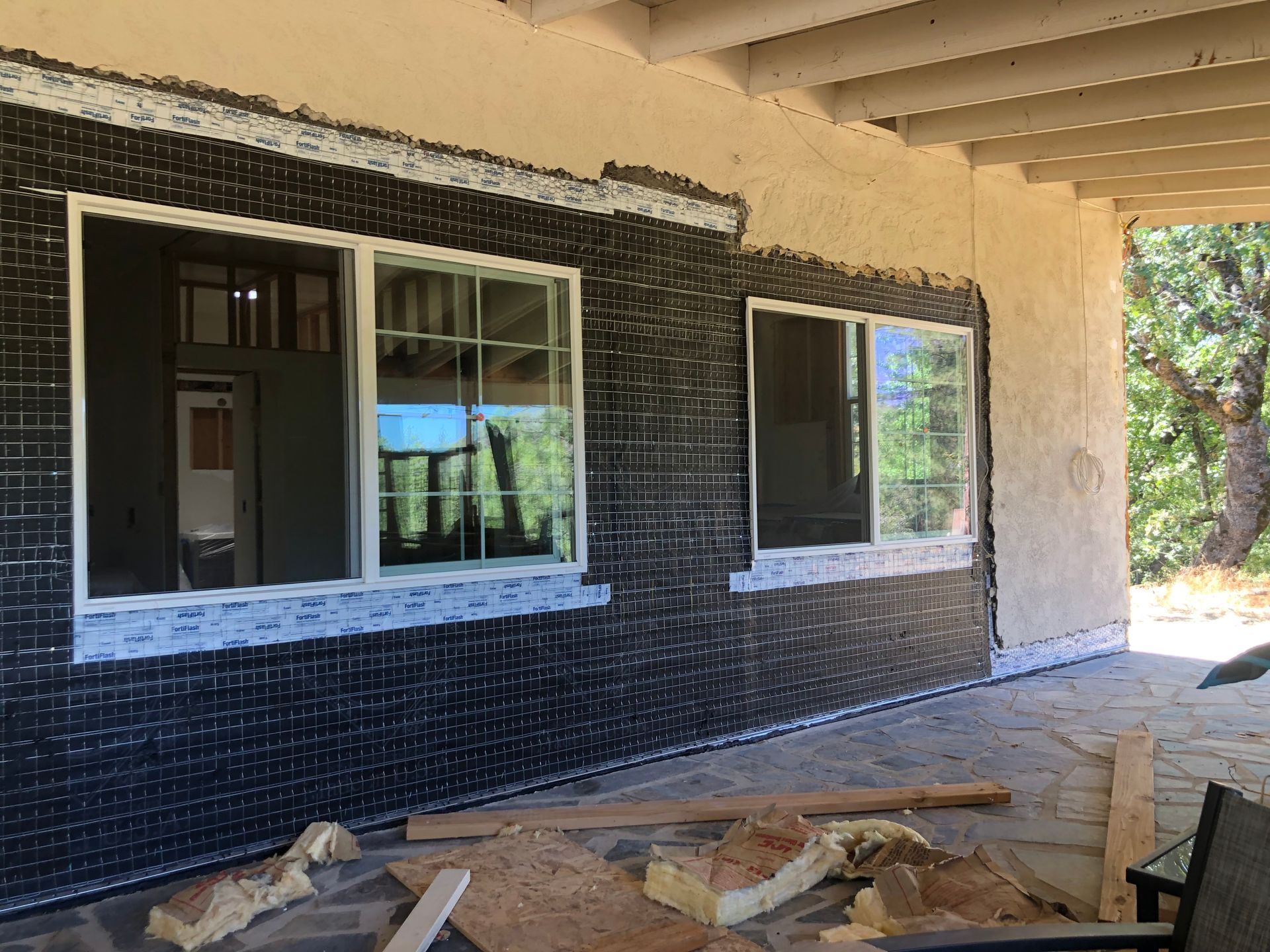 Construction site showing a house exterior with exposed black vapor barrier and new window frames under a patio cover.