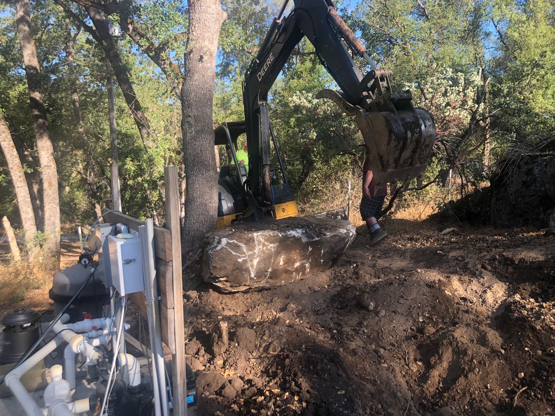 A yellow excavator operates in a wooded area, moving a large boulder near electrical equipment and a pool pump system.