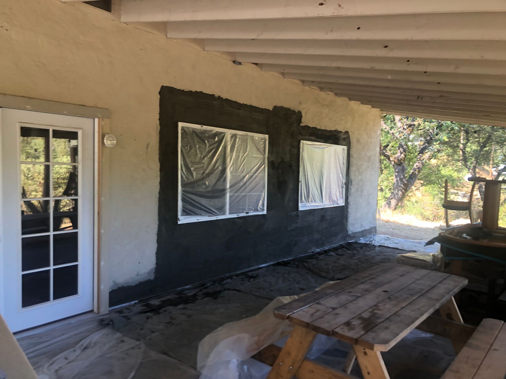 Exterior wall of a house with patches of dark gray stucco repair around two windows covered in plastic sheeting.