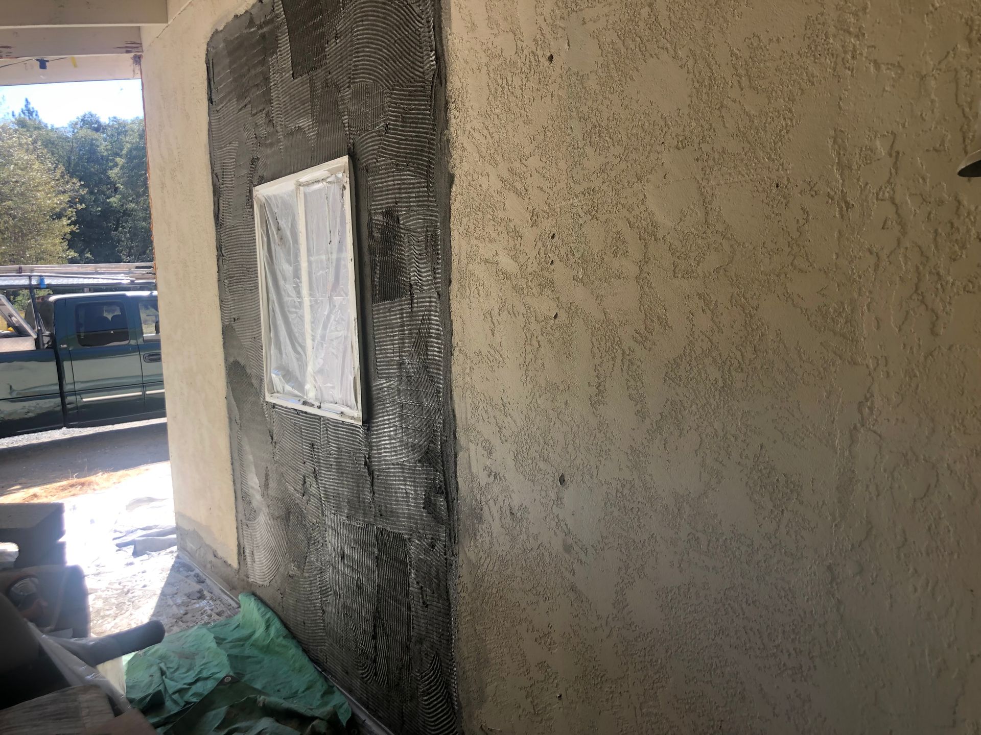 A wall under stucco repair with a rectangular window covered in plastic, showing fresh gray mortar around the frame.