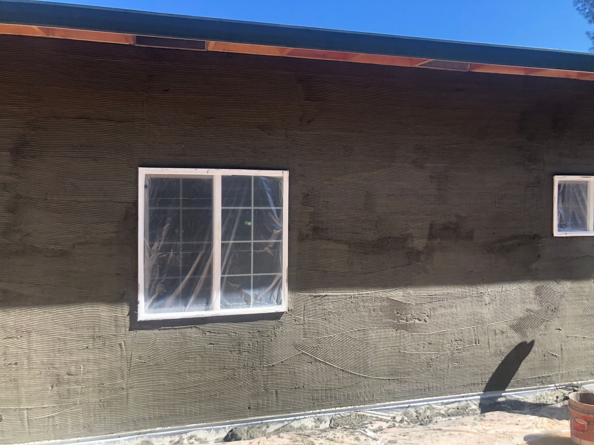 Exterior house wall under construction with a fresh coat of wet, horizontally-ridged brown stucco around white windows.