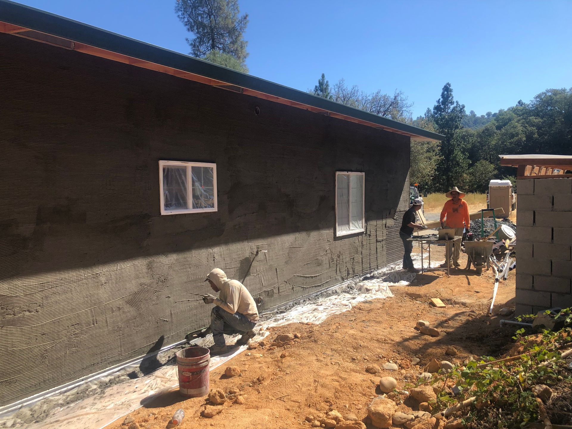 Construction workers apply stucco to the exterior wall of a building on a sunny day.