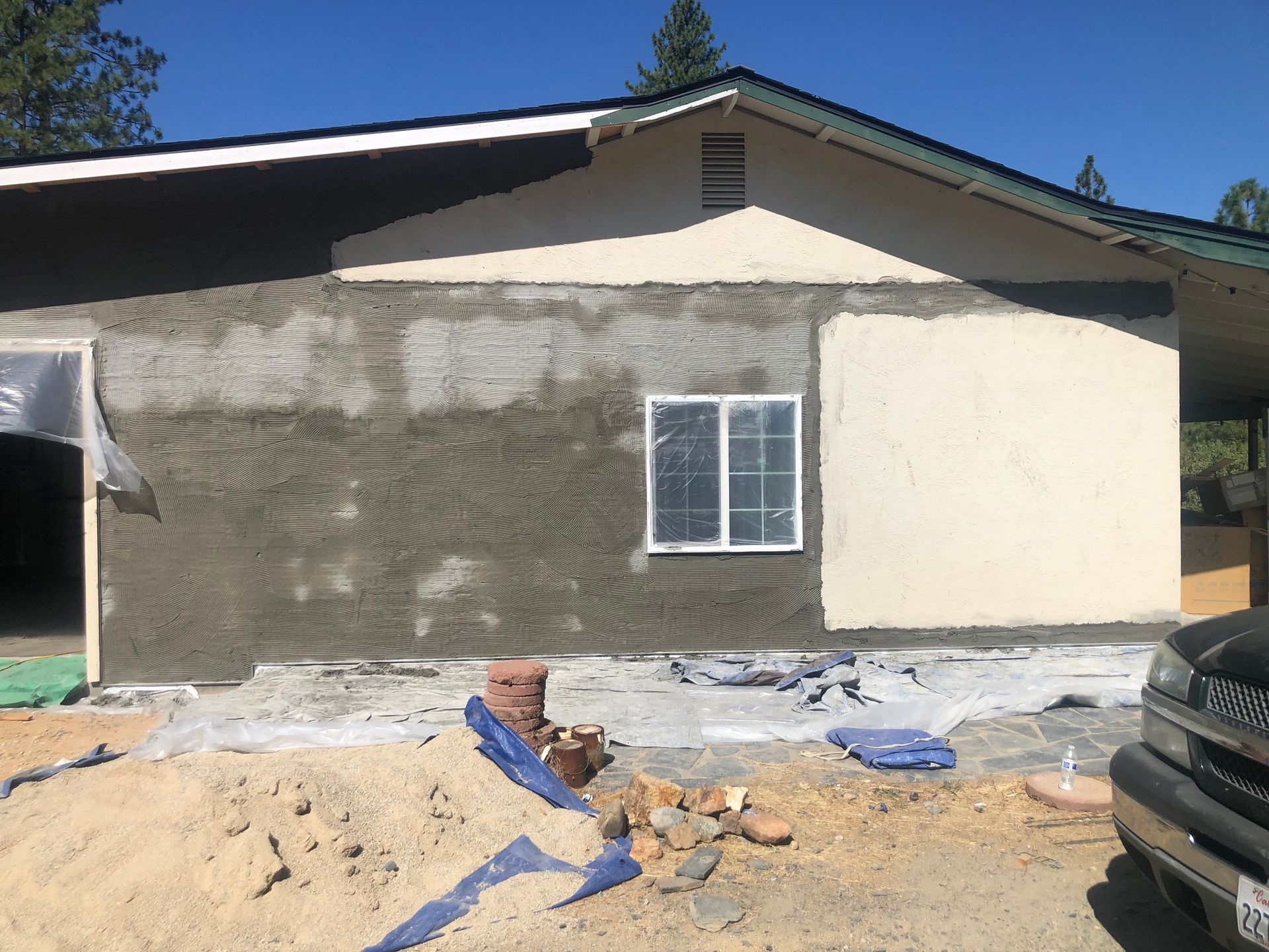 A house exterior under construction with partial stucco siding, a window, and sand scattered on the ground in front.
