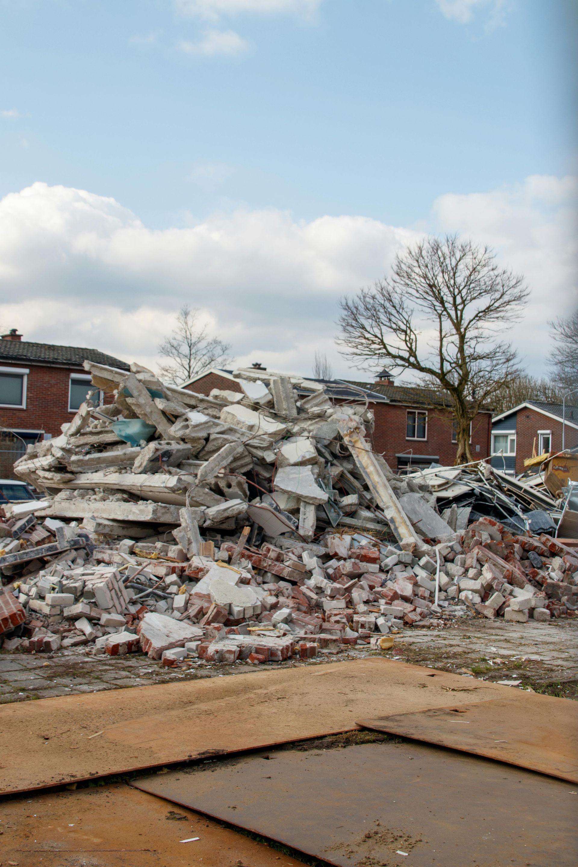 A large pile of concrete rubble and broken bricks from a demolished building sits outdoors near houses and a bare tree.