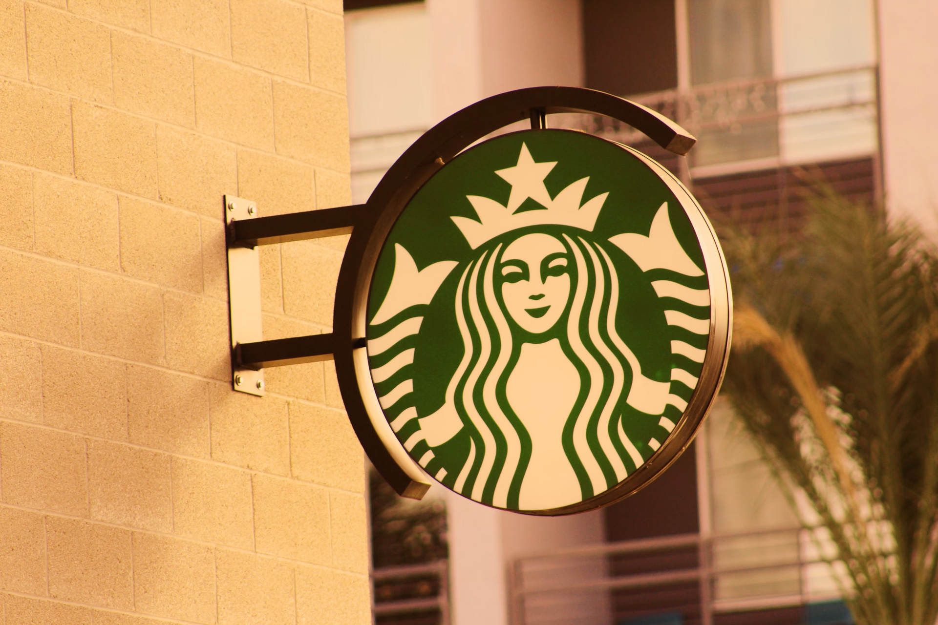 Starbucks sign, green and white logo, hanging on tan brick wall.