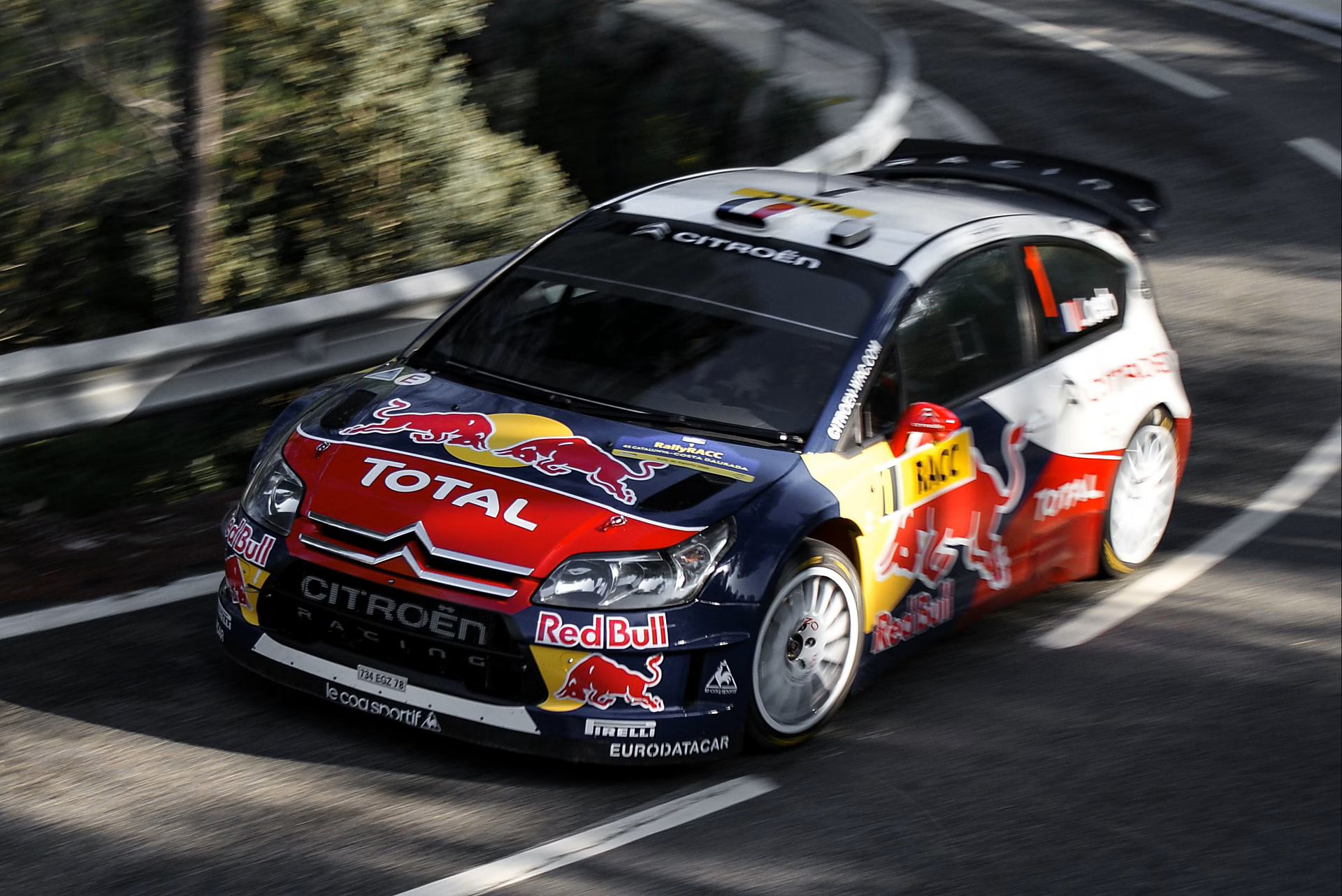 Citroen rally car with Red Bull and Total logos speeding on a curved road, trees in the background.